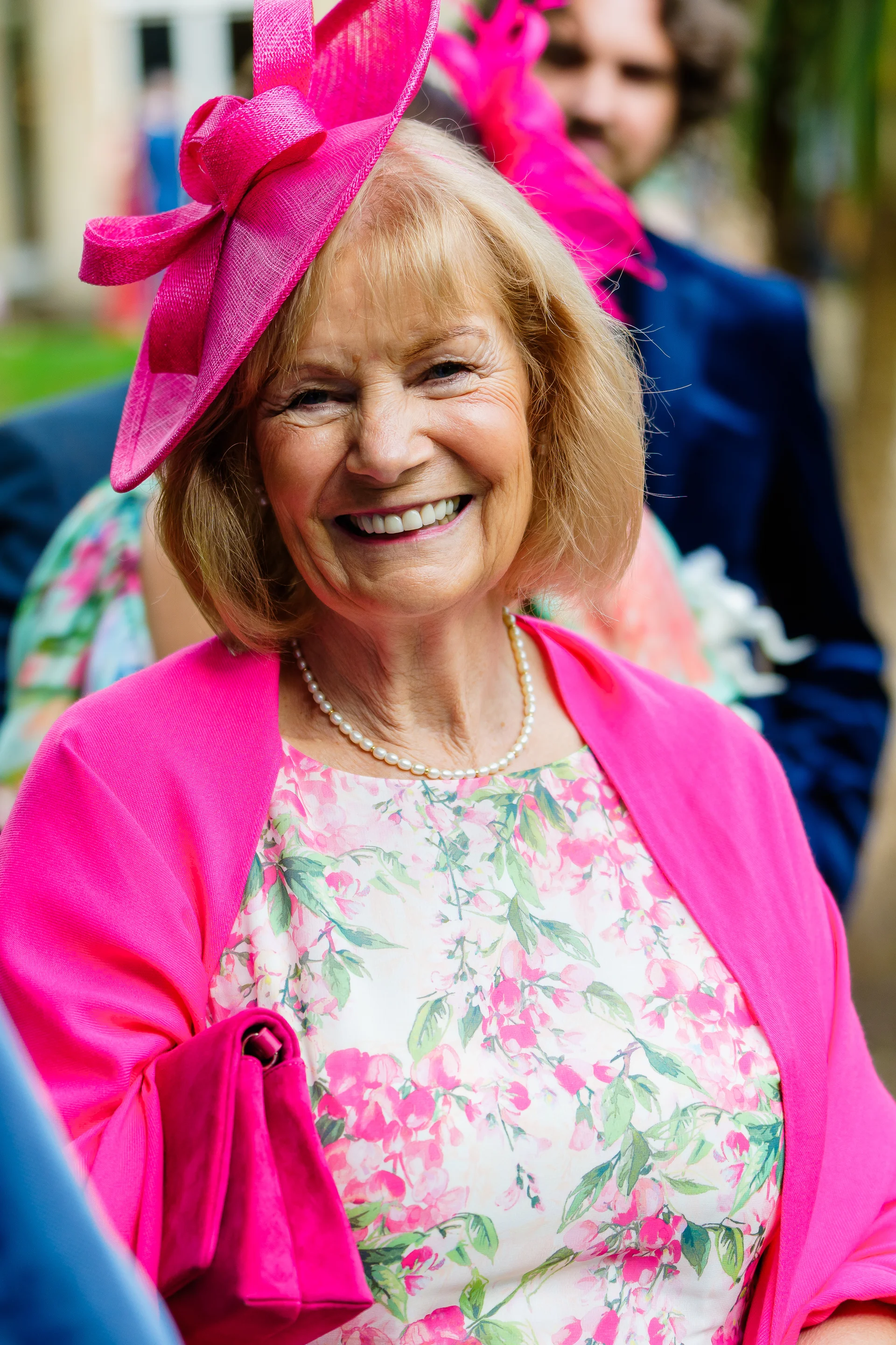 An elderly female wedding guest wearing a bright pink fascinator, floral dress, and pearl necklace smiles warmly outdoors during the celebration.