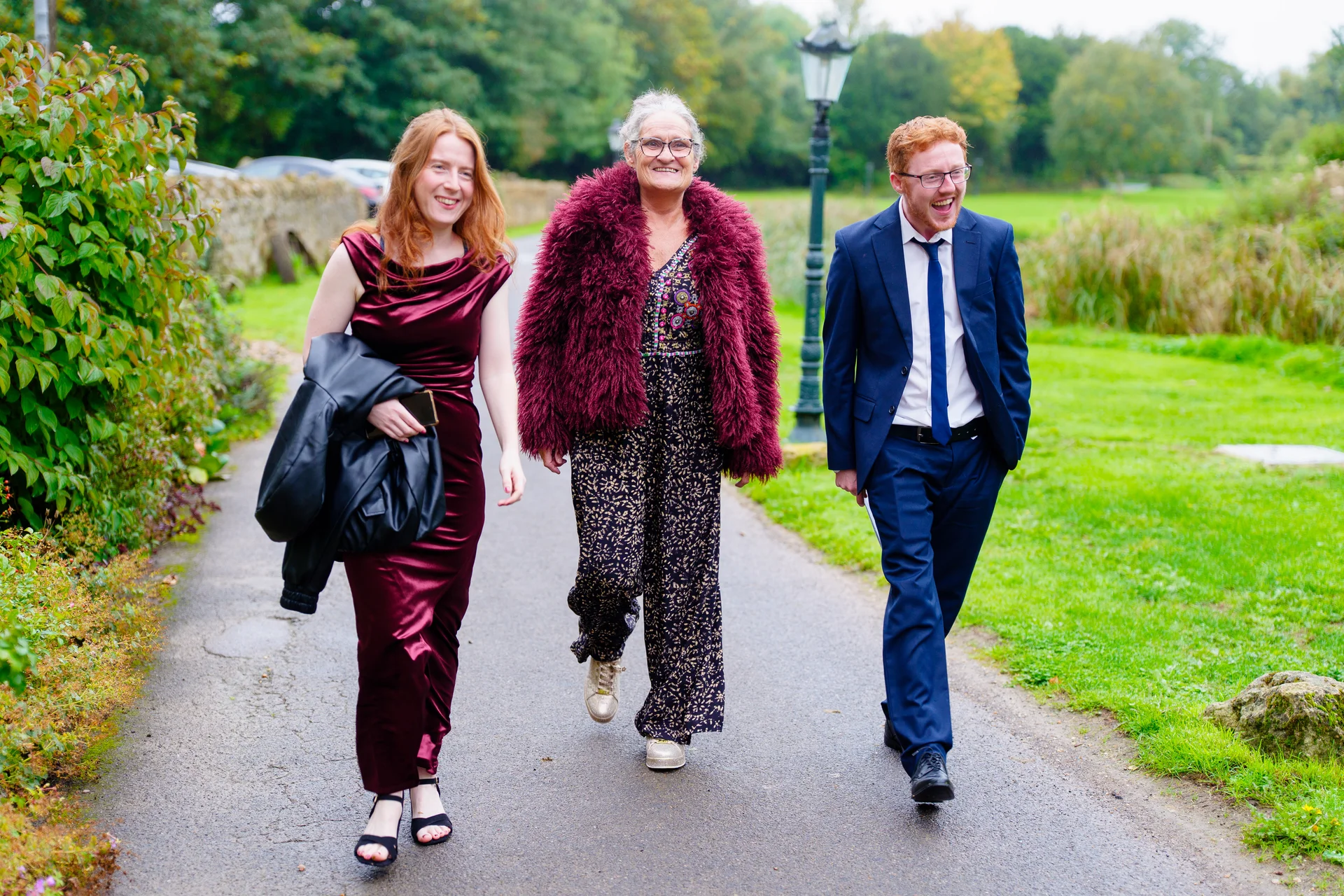 Three wedding guests laughing and walking along a paved garden pathway surrounded by lush green lawns and trees.