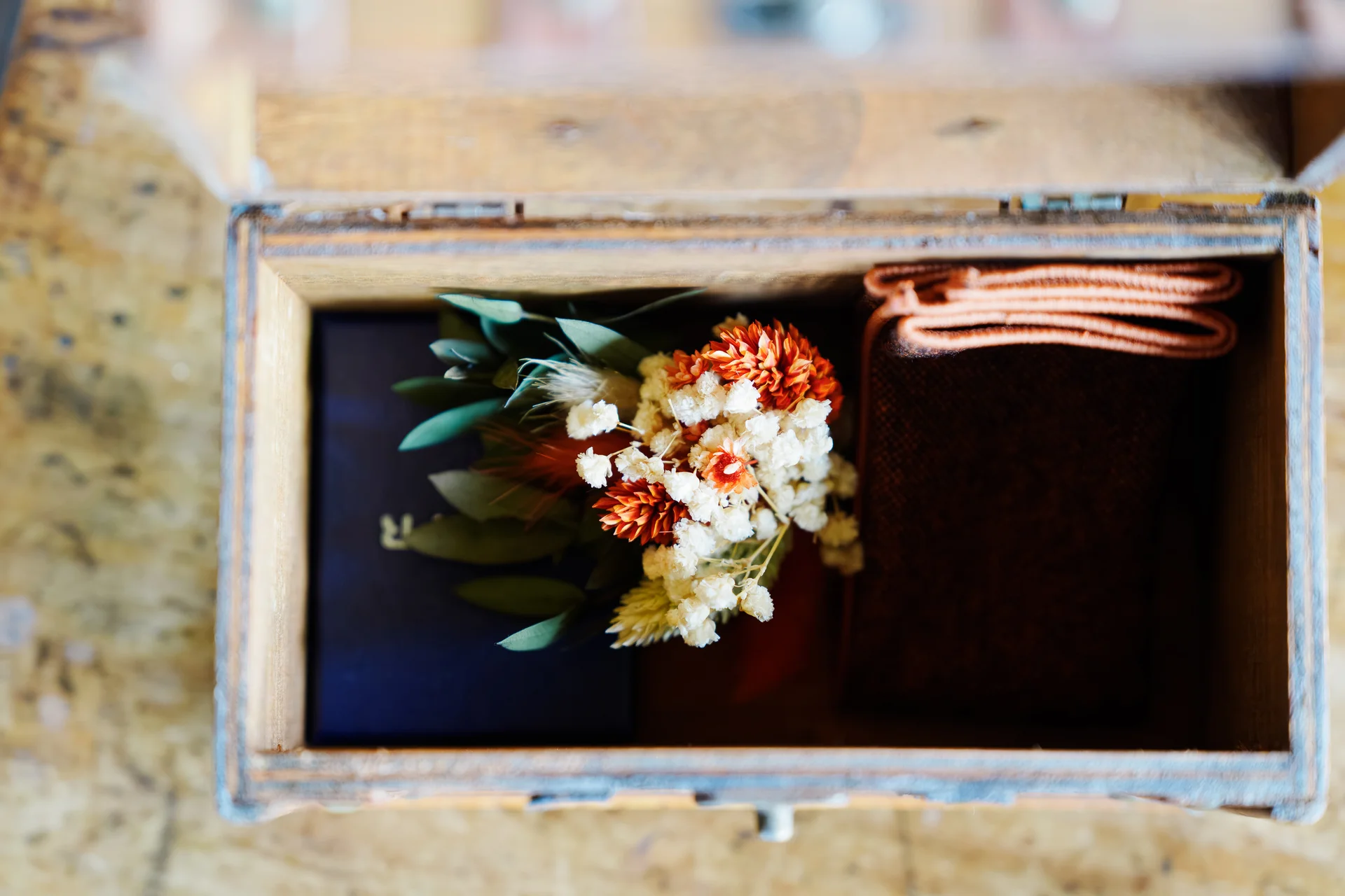 An overhead macro shot of a small wooden memory box holding a dried floral buttonhole alongside folded fabric details.