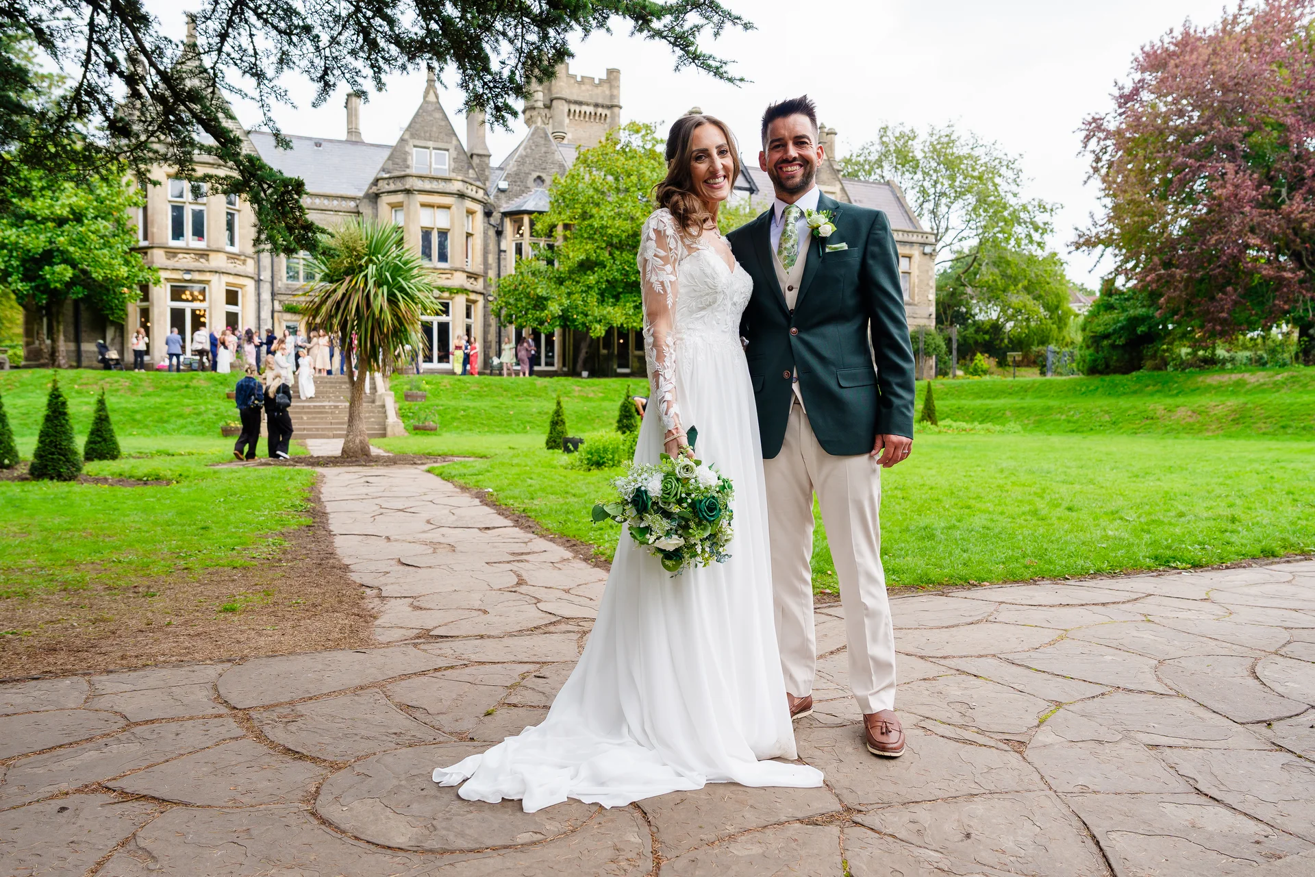 A smiling bride and groom stand on a stone patio in front of a grand manor house, holding a bouquet with white and green florals.