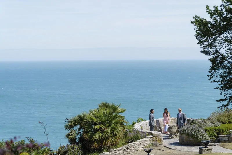 A wide angle view of guests standing on a sunny clifftop terrace overlooking an expansive blue sea and horizon.