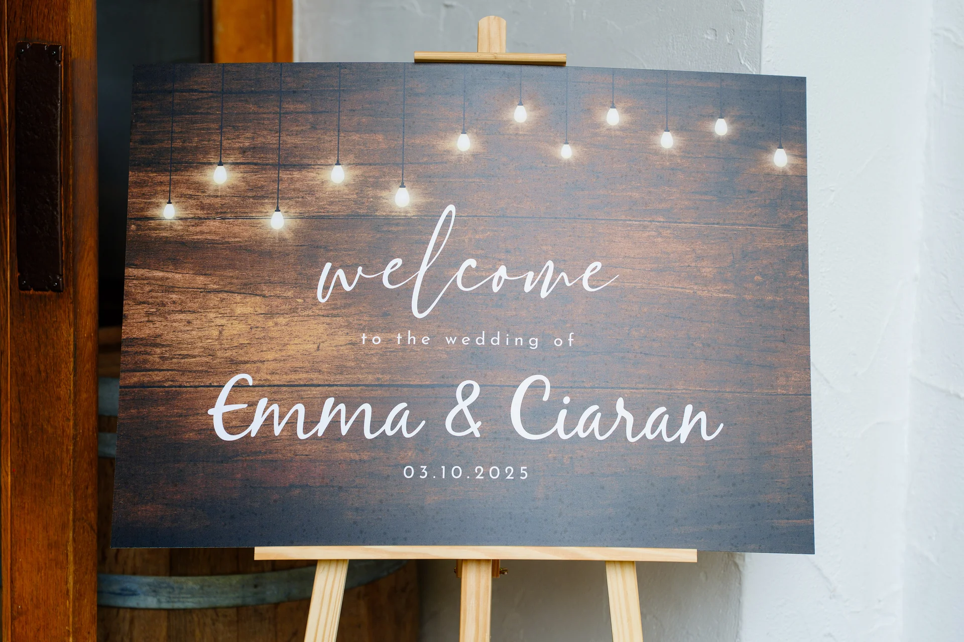 A rustic wooden-textured wedding welcome sign featuring fairy lights and white script text displaying the names Emma and Ciaran on a wooden easel.