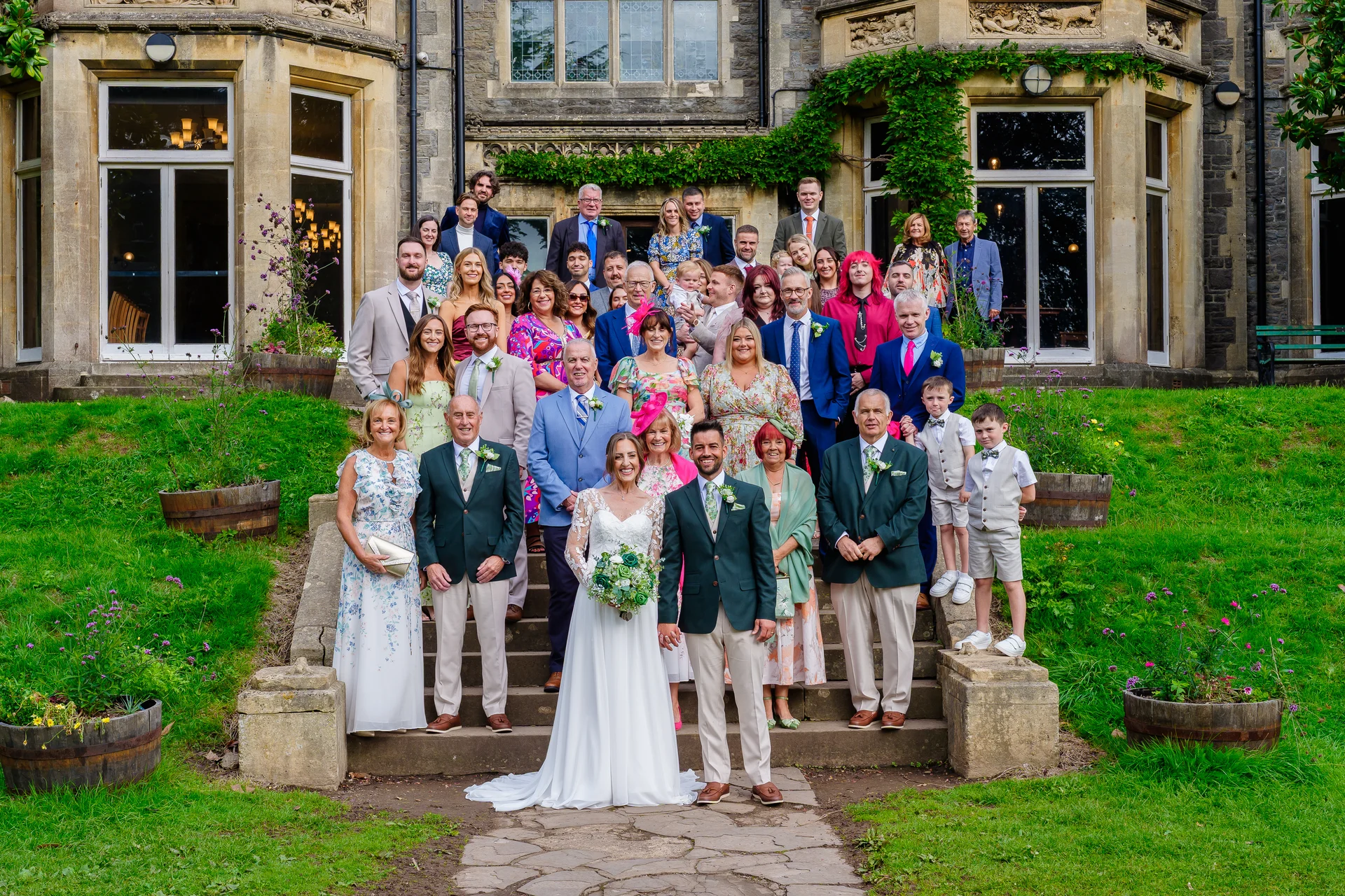 The bride and groom pose with their large wedding party and guests on the grand stone steps outside a stunning country house venue during a summer celebration.
