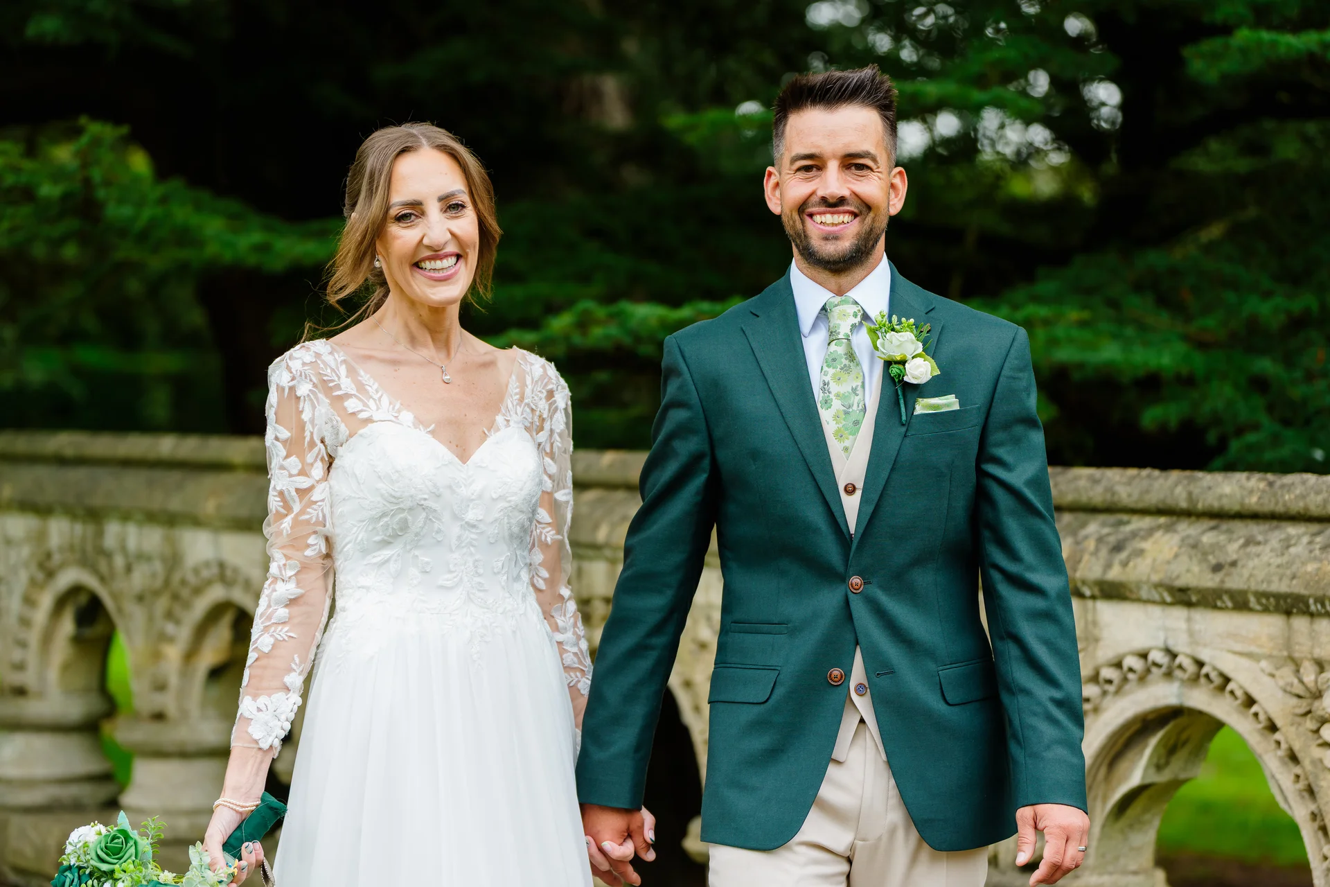 A smiling bride in a lace-sleeved a-line gown and a groom in a dark green suit walk hand-in-hand outdoors near a stone bridge.