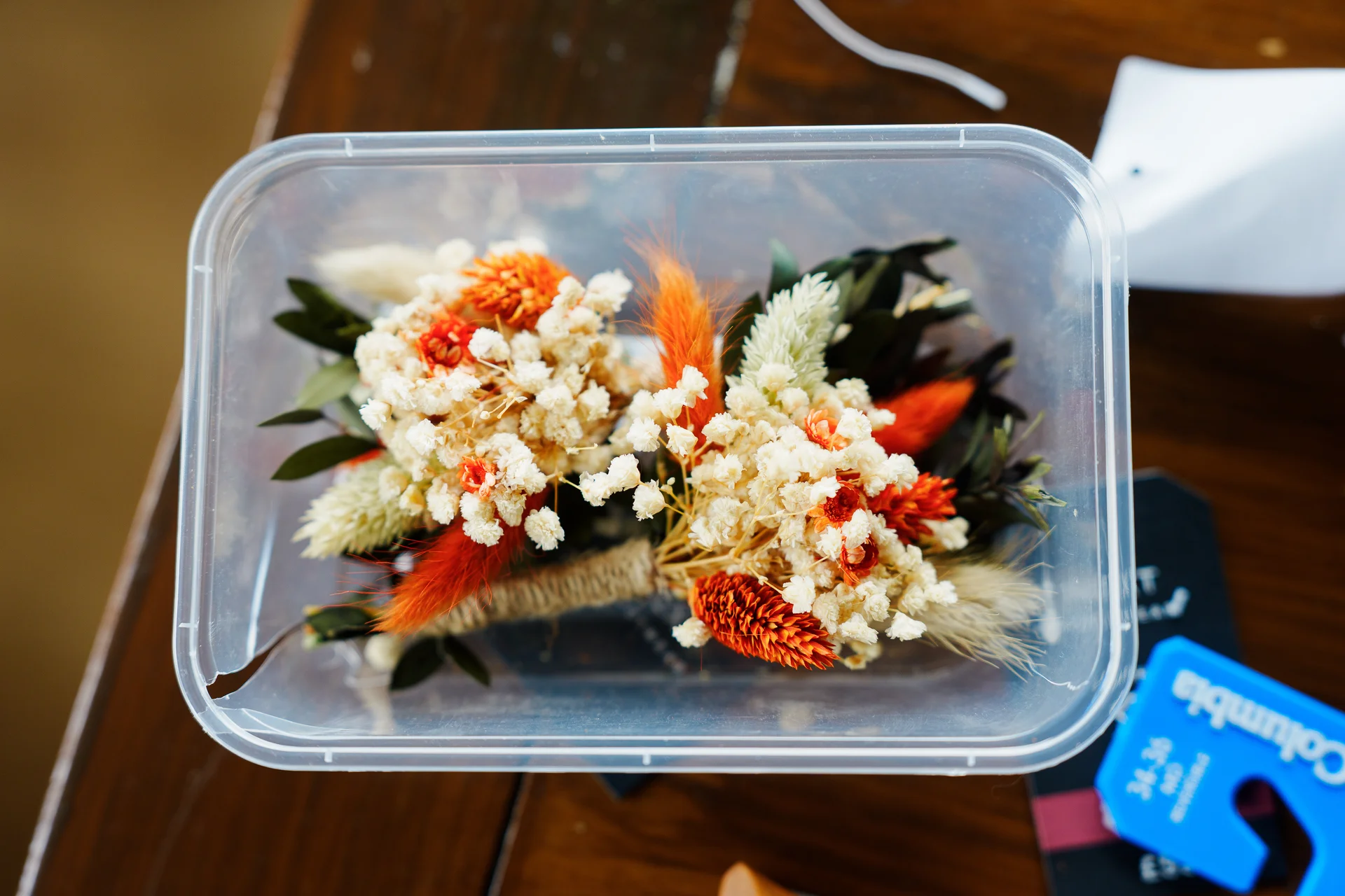 A handmade dried flower buttonhole with burnt orange and cream elements rests inside a clear plastic storage container on a wooden table.