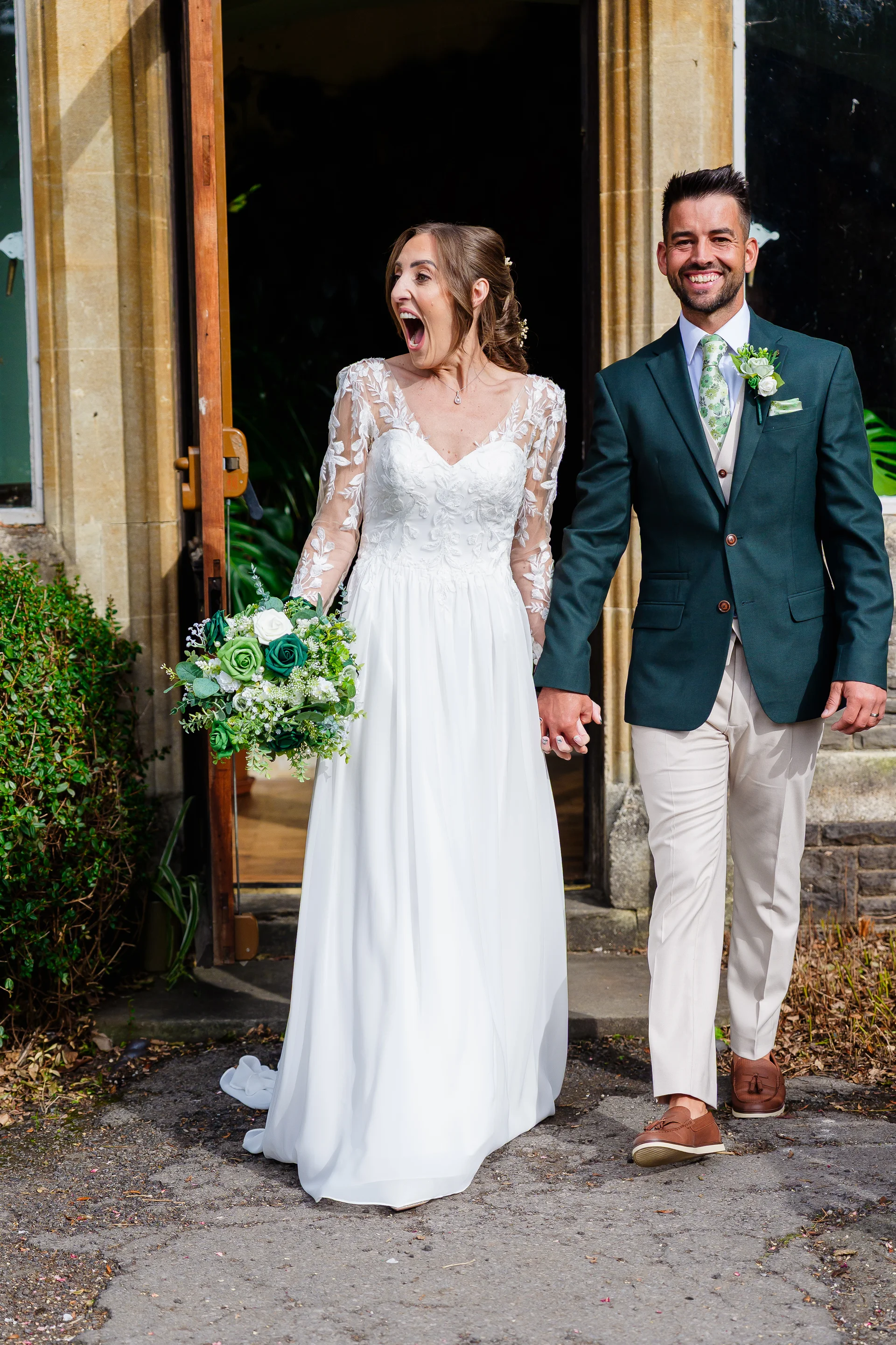 A newlywed couple excitedly emerges from a venue doorway holding hands, with the bride laughing joyfully and holding a green-themed bouquet.
