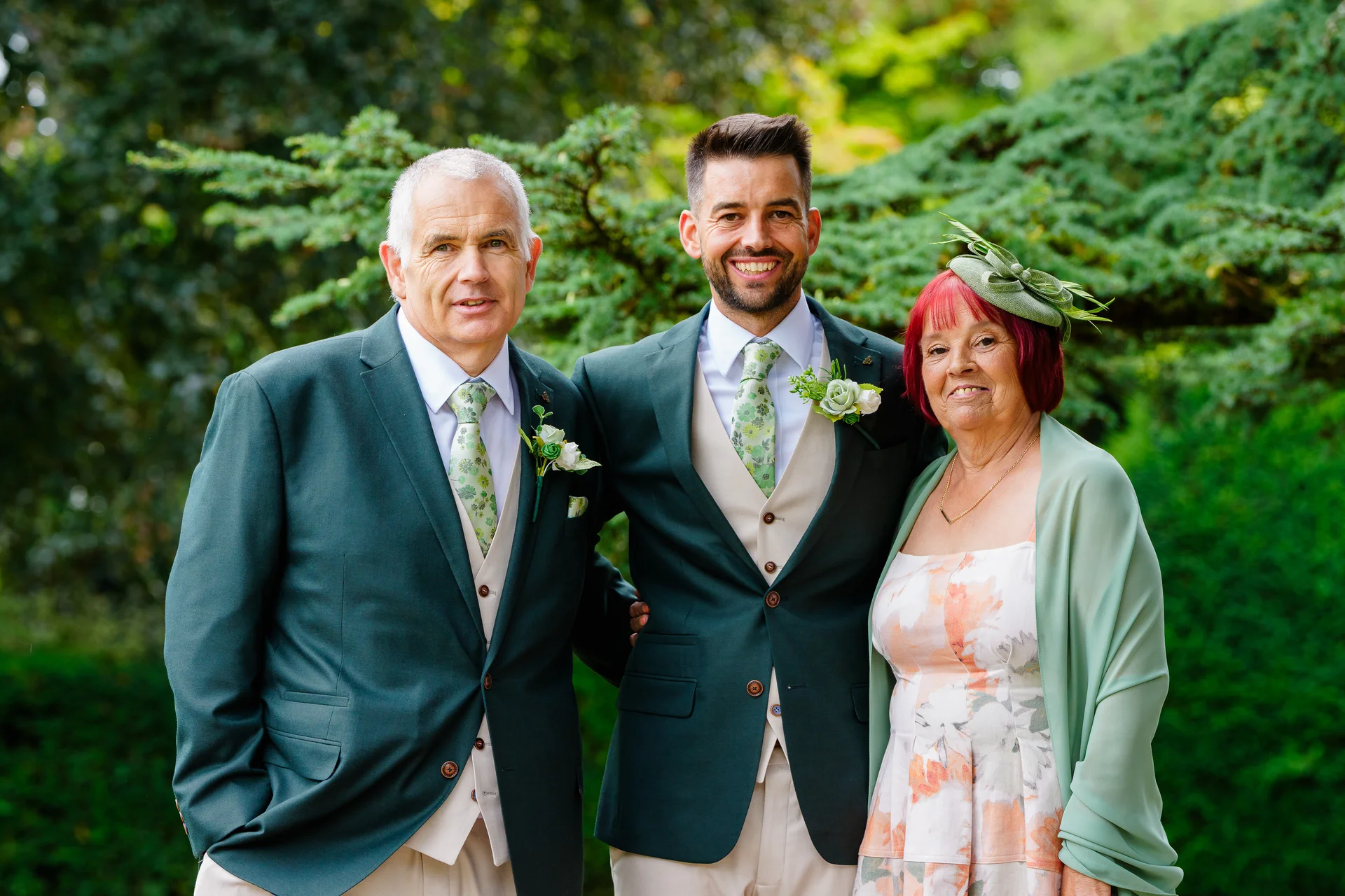 A smiling groom in a dark green suit stands outdoors flanked by his parents, with his mother wearing a floral dress, sage shawl, and matching fascinator.