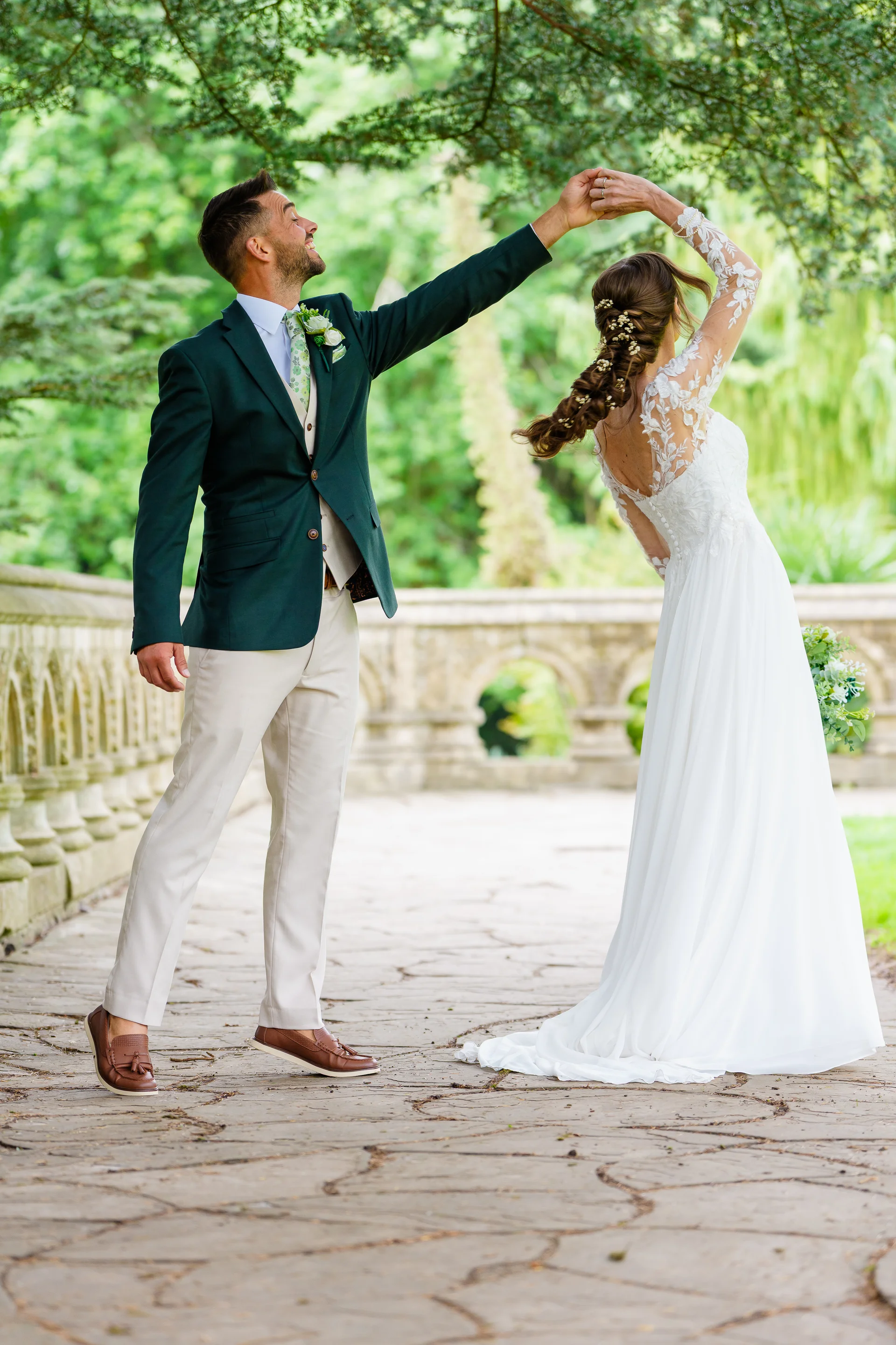 A groom in a green suit and a bride in a lace-sleeved gown dance together outdoors on a stone bridge framed by lush greenery.
