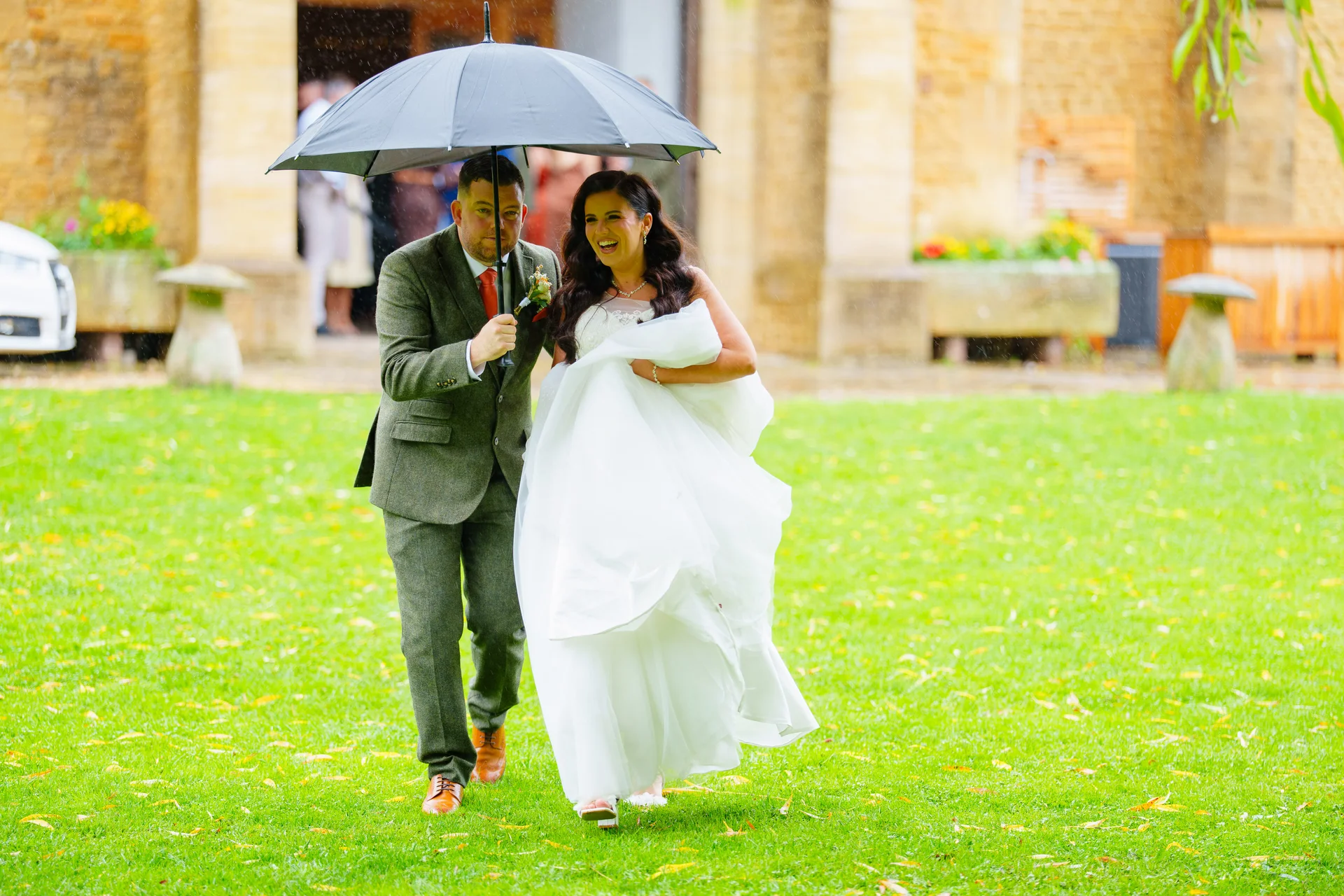 A newlywed couple laugh joyfully as they walk across a lush green lawn together under a single black umbrella during a rainy wedding day.