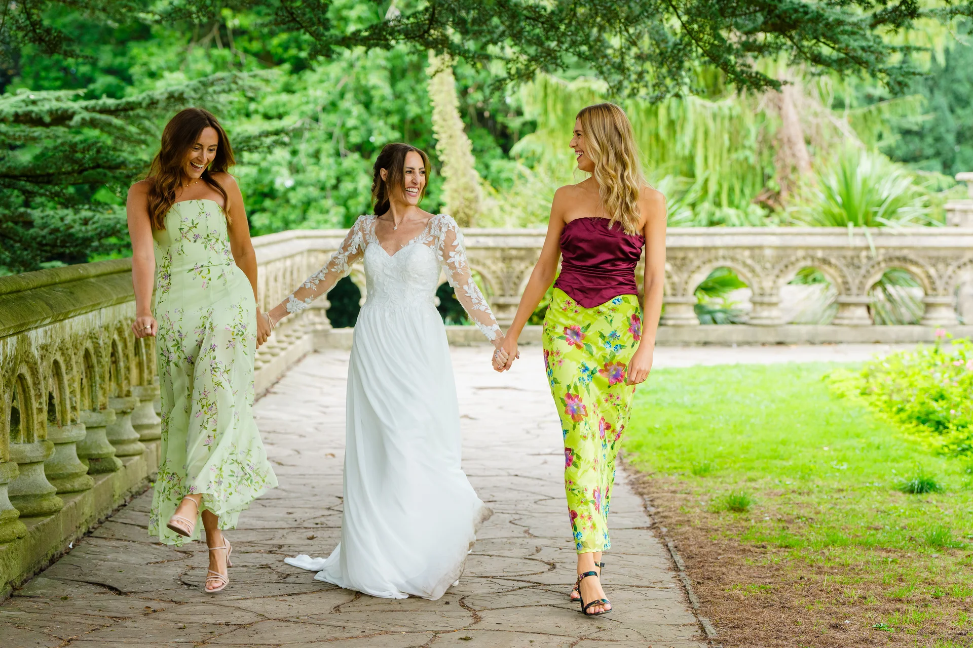 A bride in a long-sleeved lace wedding gown walks hand in hand along a stone path with two bridesmaids in mismatched floral dresses, laughing joyfully outdoors.