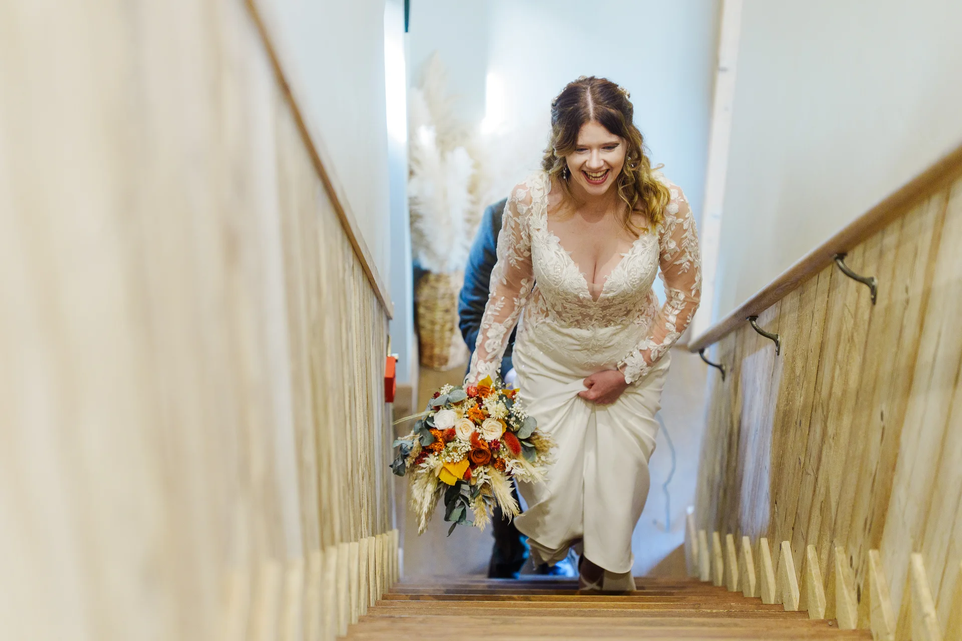 A joyful bride laughs while walking down a wooden staircase holding an autumnal bouquet with burnt orange and white flowers.