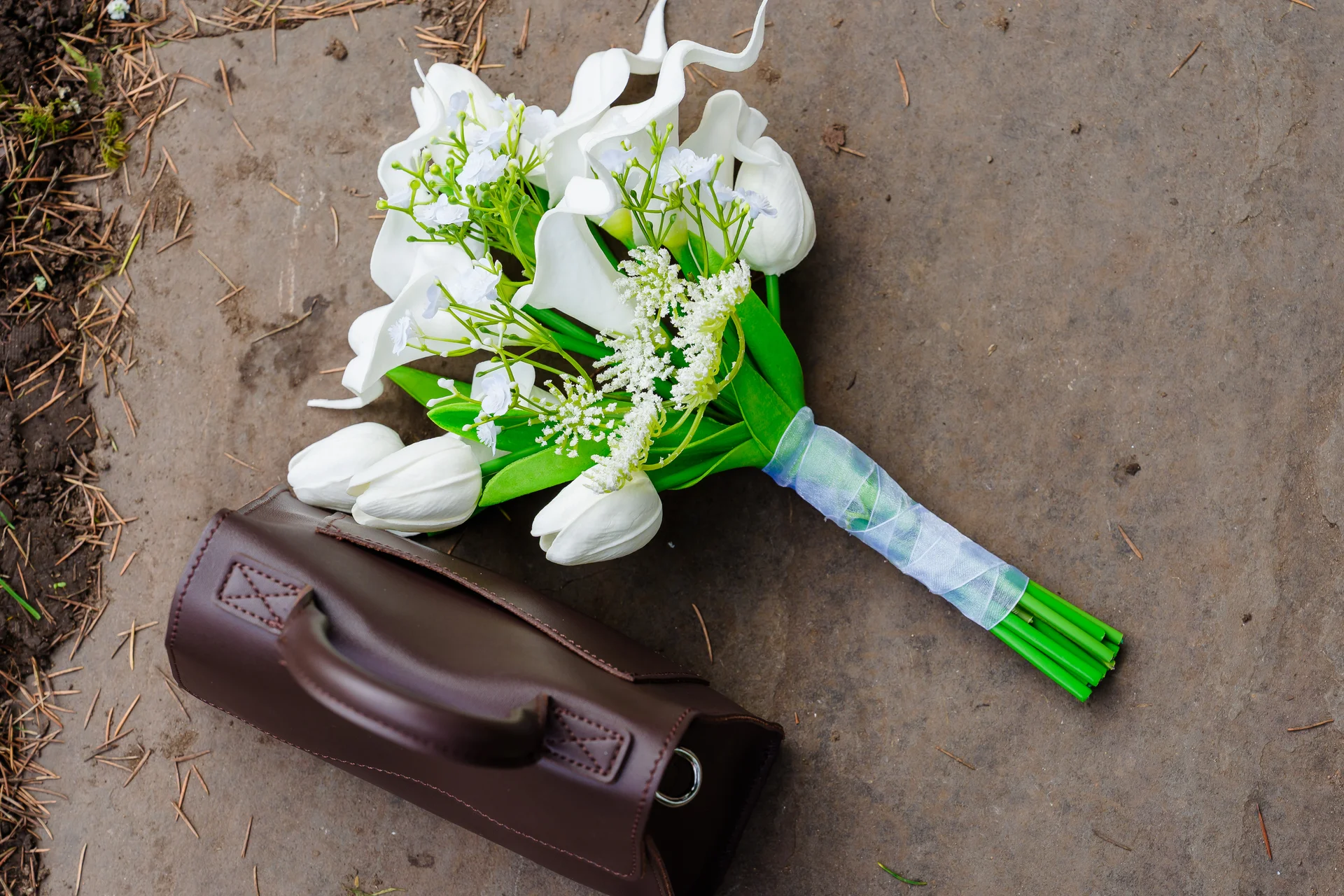 An overhead flat lay shot of a white hand-tied bridal bouquet featuring calla lilies and roses, resting on a stone surface beside a brown leather bag.