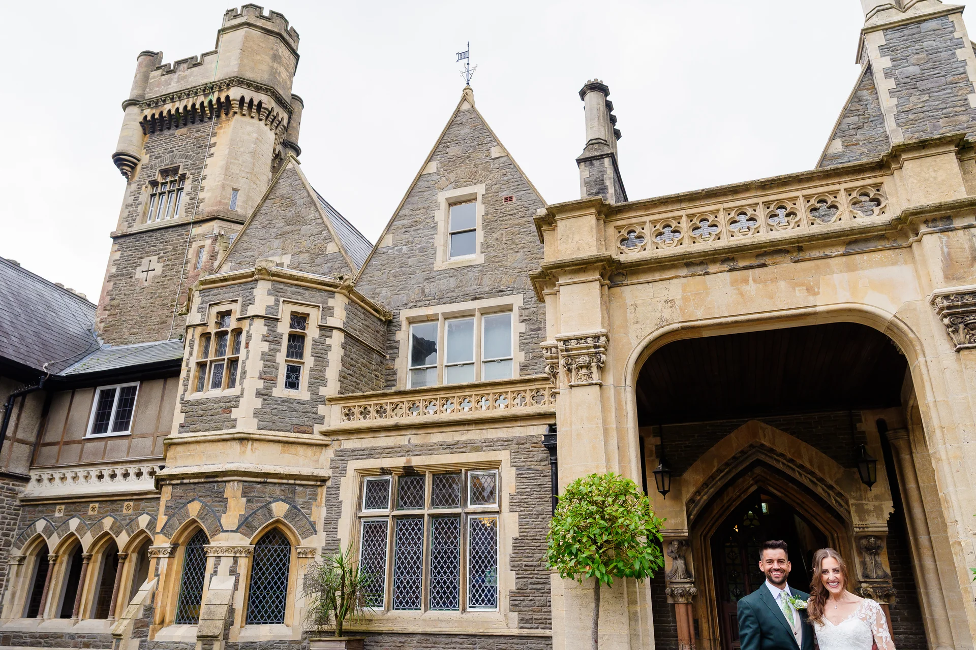 A bride and groom smiling happily together in front of the grand stone facade and arched entrance of a historic castle wedding venue.
