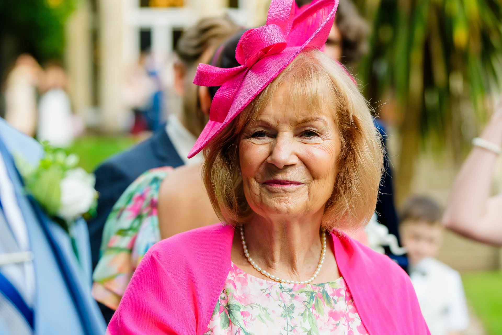 An elderly female wedding guest wearing a bright pink fascinator and matching jacket smiling warmly outdoors during the reception.