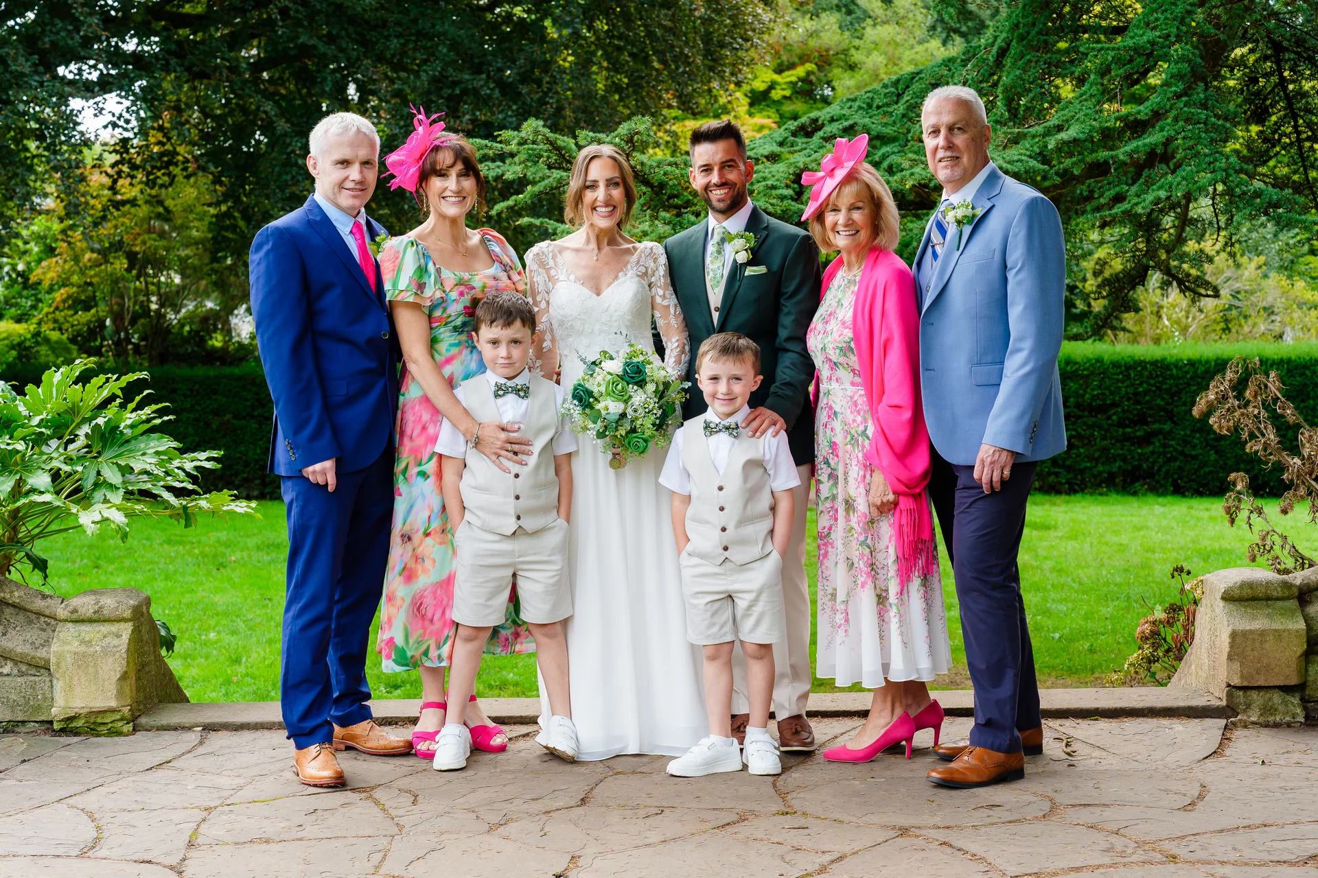 A happy bride and groom pose outdoors in a lush green garden alongside family members, including page boys in waistcoats and guests wearing bright pink fascinators.