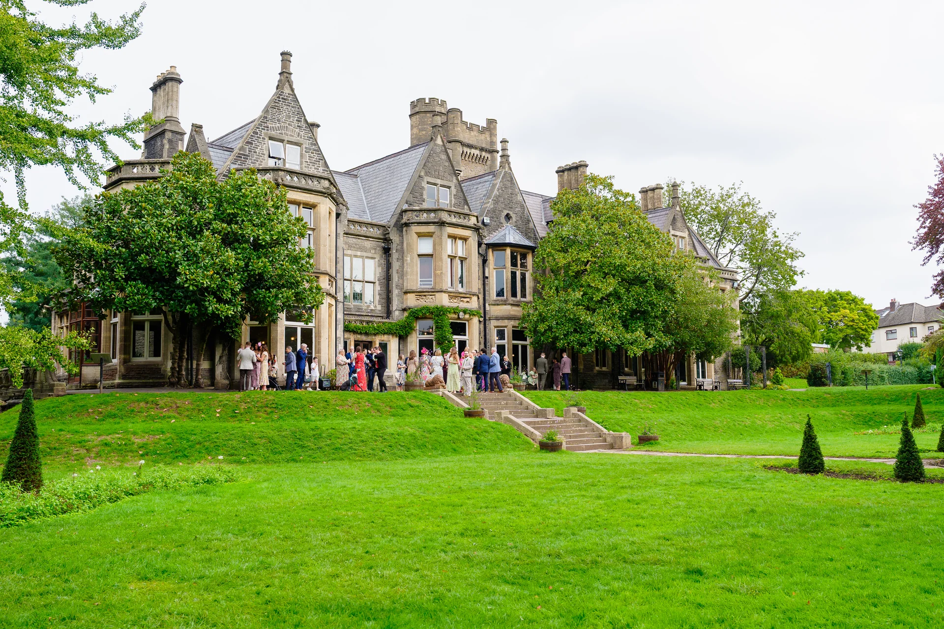 Wedding guests gather outside a grand historic country house venue during a sunny drinks reception on the sweeping lawn.