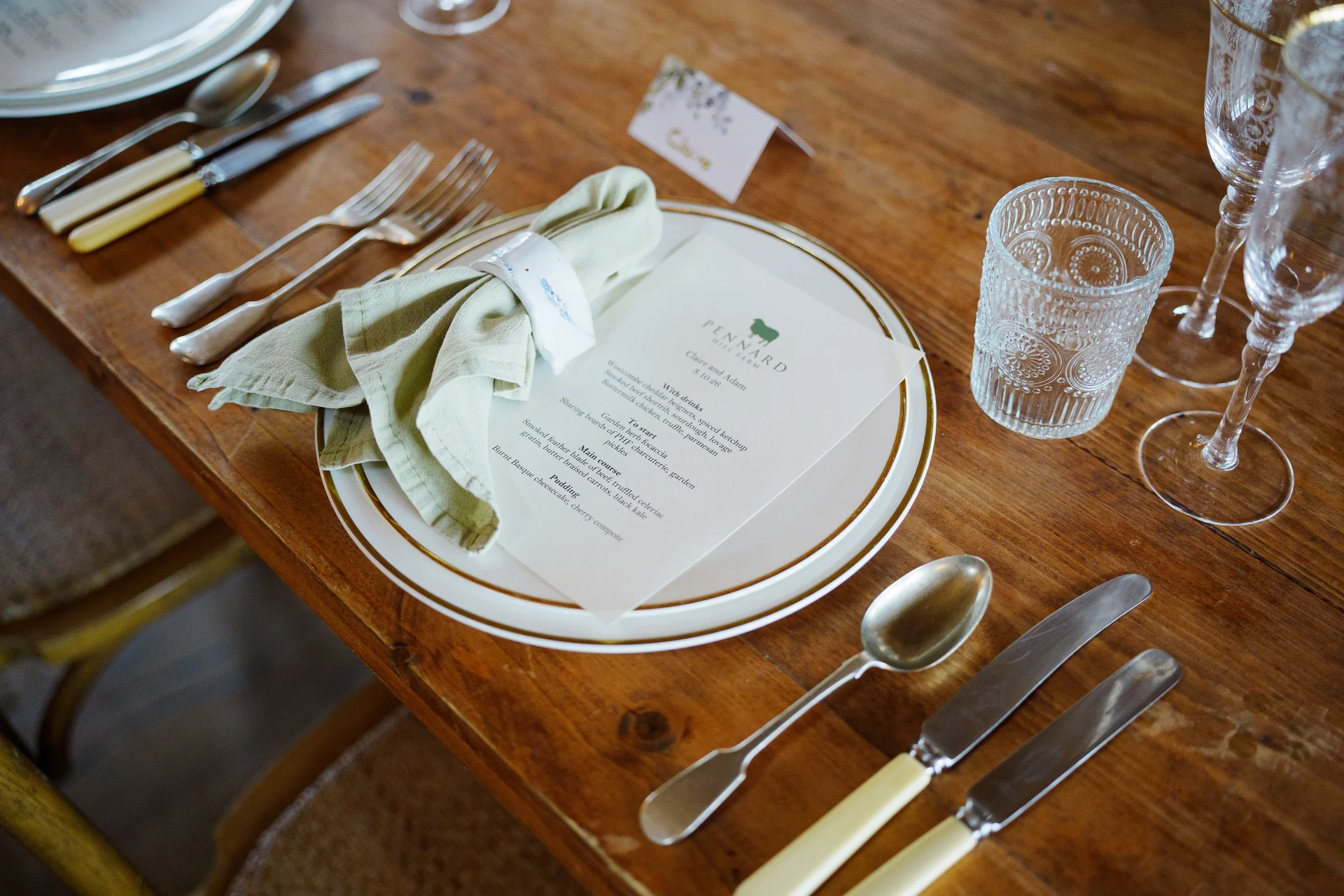 A beautifully styled rustic wedding place setting features a layered white plate, an elegant menu, a sage green napkin with a ceramic ring, and vintage glassware on a wooden table.