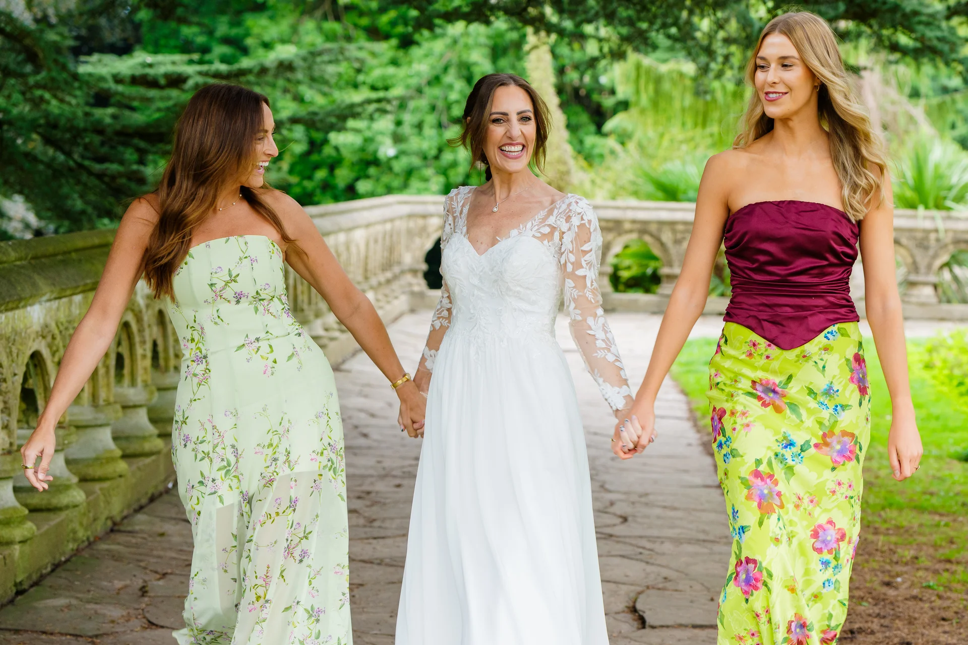 A joyful bride in a long-sleeved lace gown walks along a stone pathway, holding hands and laughing with her two bridesmaids wearing mismatched floral dresses.