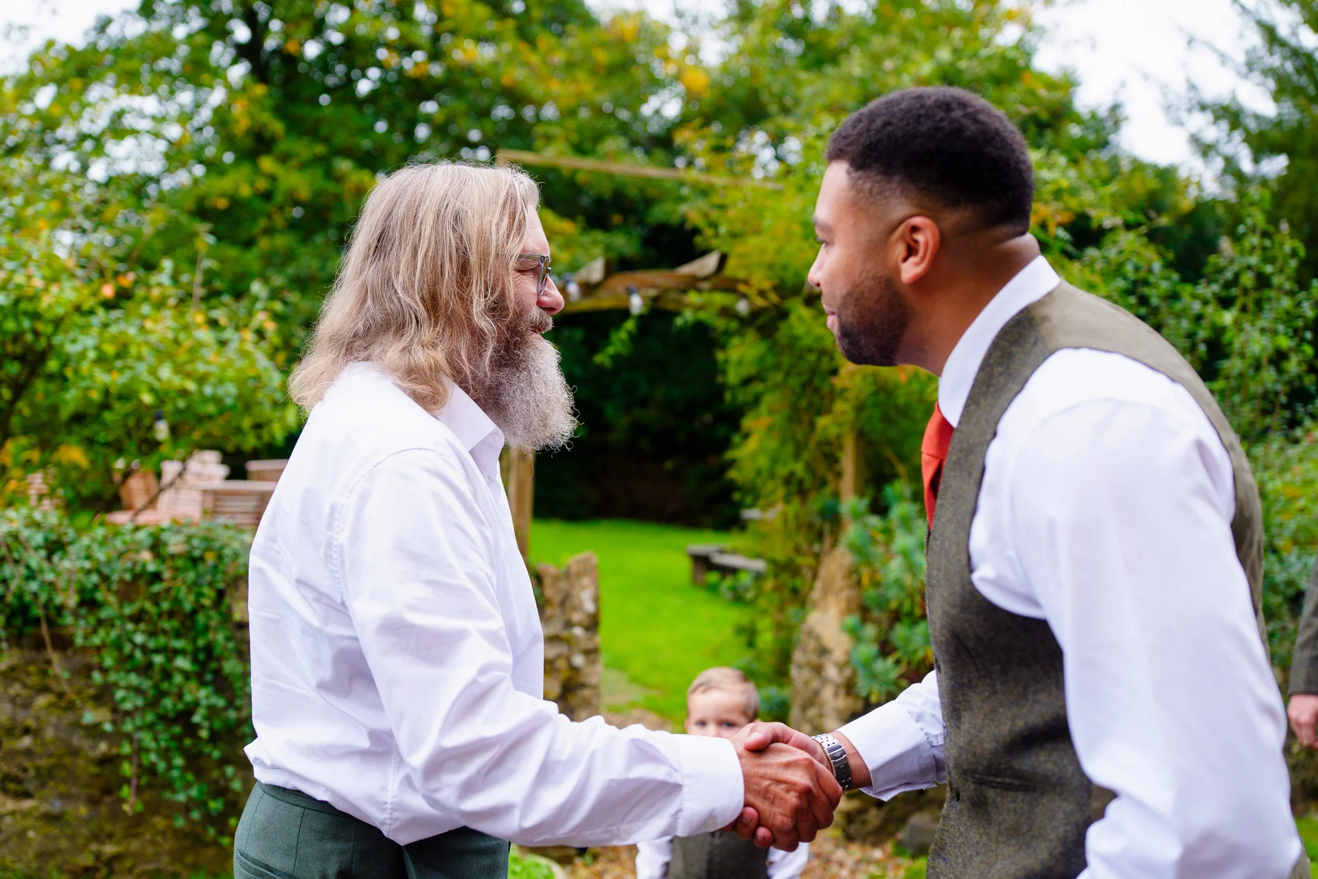 Two grooms share a warm handshake outdoors in a lush garden setting during their wedding celebration.
