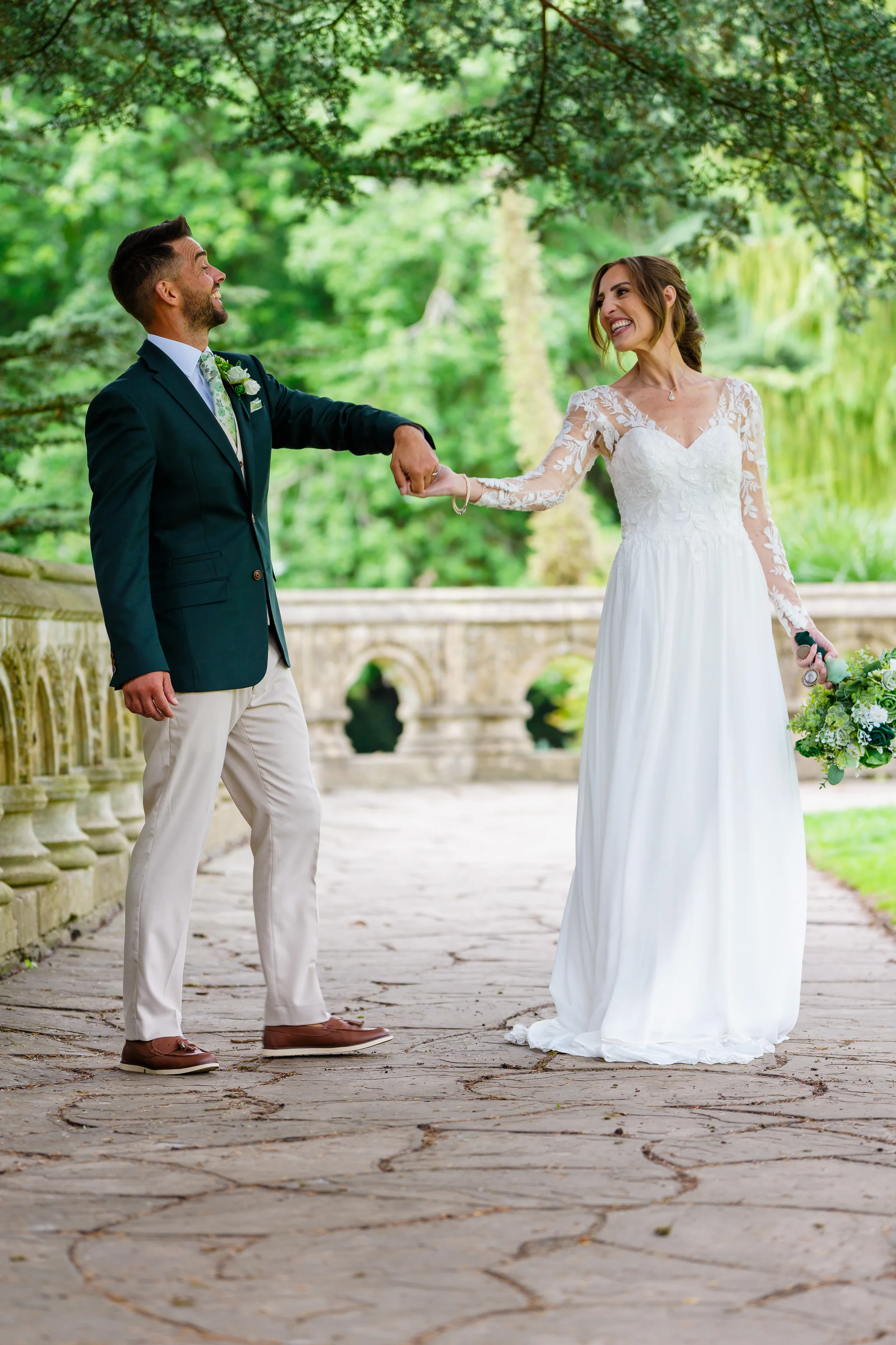 A joyful bride and groom laugh together while holding hands on a stone bridge surrounded by lush green foliage on their wedding day.