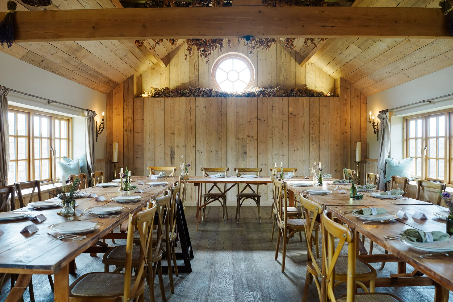 A sunlit rustic barn wedding venue features long wooden banquet tables arranged neatly with place settings, tapered candles, and cross-back chairs under a vaulted wooden ceiling.