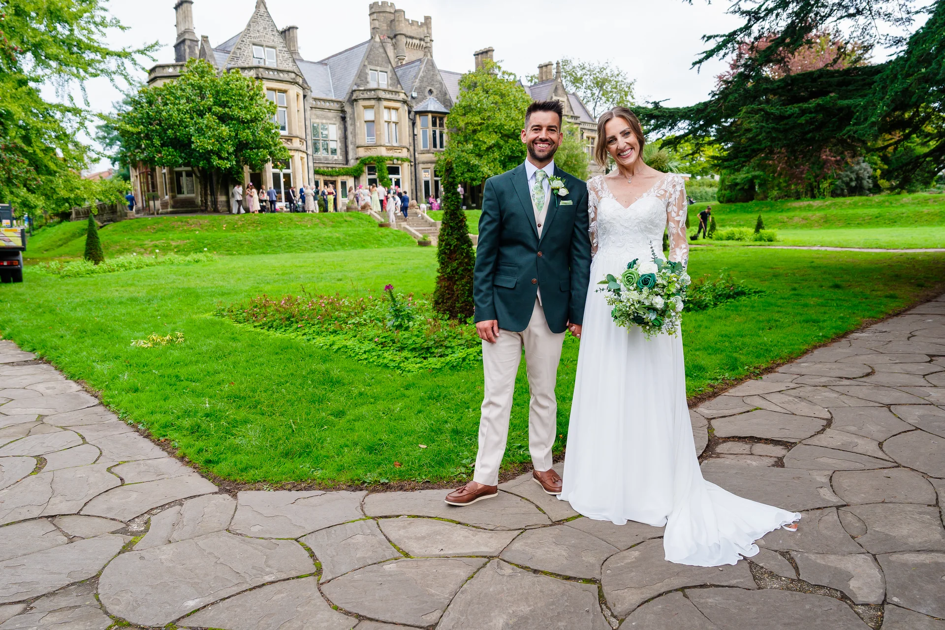 A smiling bride and groom stand outdoors on a stone pathway in front of a grand country house, with the bride holding a lush green and white bouquet.
