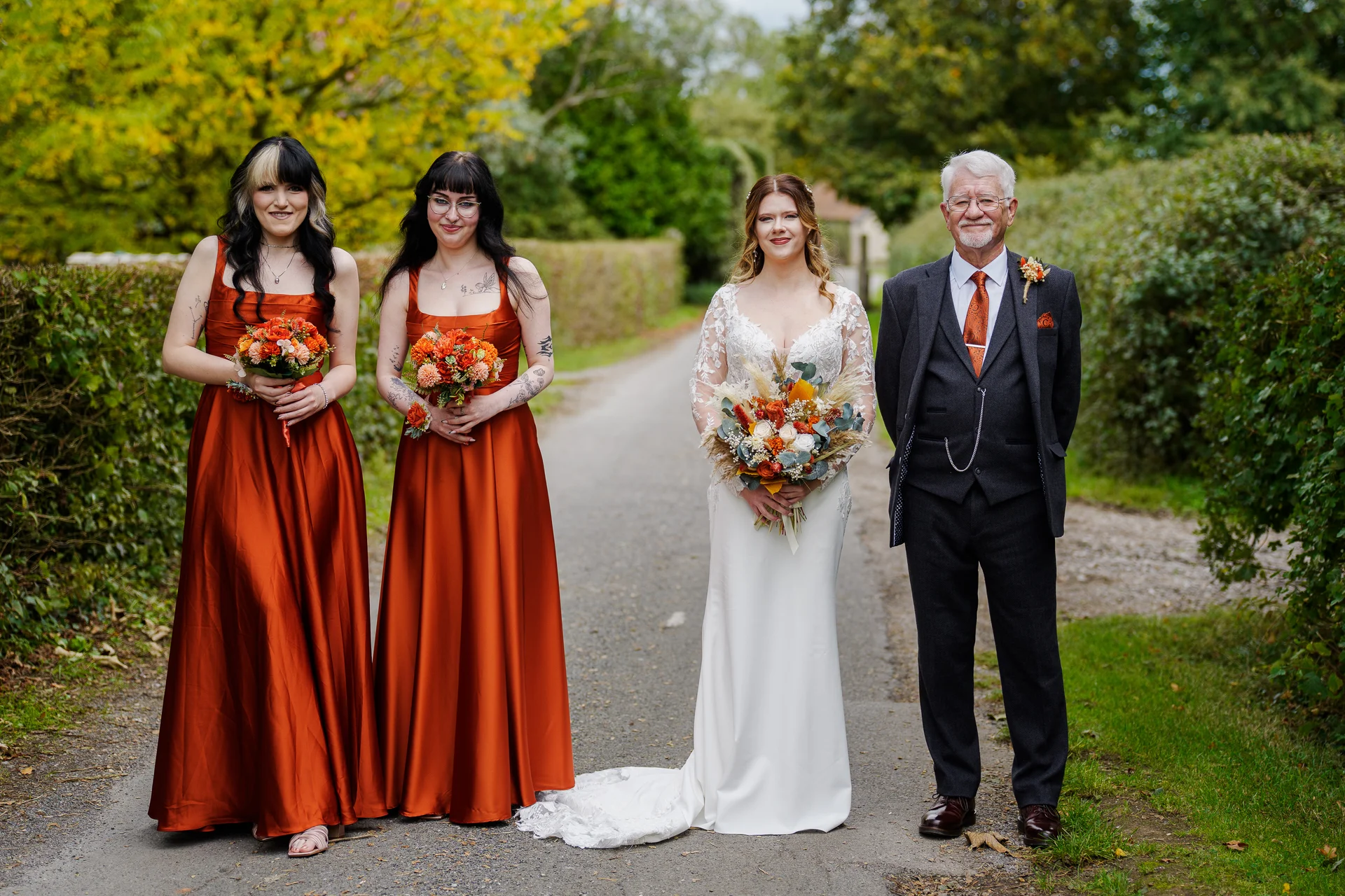 A bride in a long-sleeved white crepe gown stands on a country lane holding a vibrant autumn bouquet, flanked by two bridesmaids in rust-orange satin dresses and a gentleman in a charcoal three-piece suit.