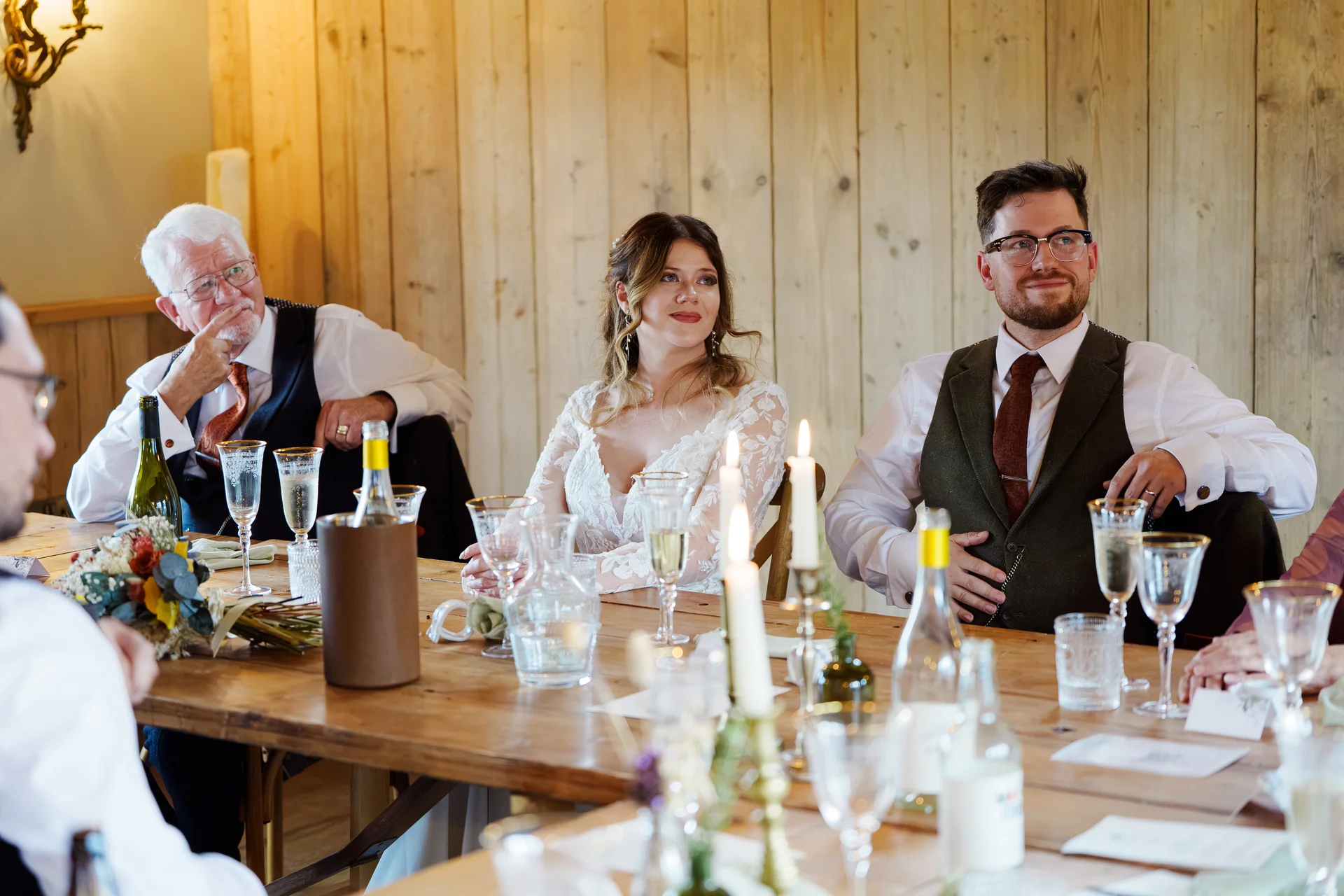 A bride, groom, and guest sit at a long wooden banquet table during wedding speeches, smiling as they listen to the toast.