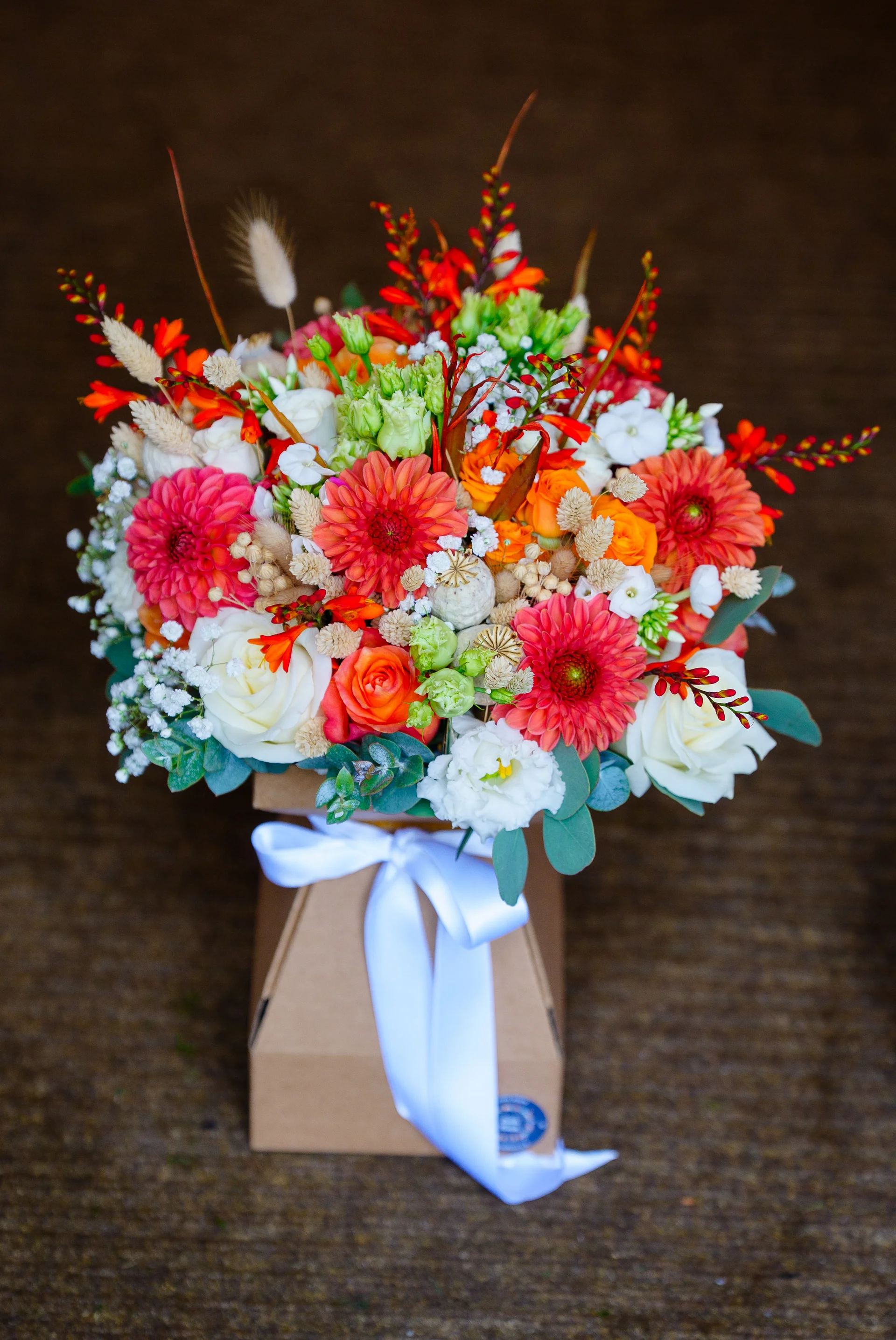 A vibrant wedding bouquet featuring vibrant orange and pink dahlias, white roses, and delicate dried grasses sits in a kraft gift box tied with a white satin bow.