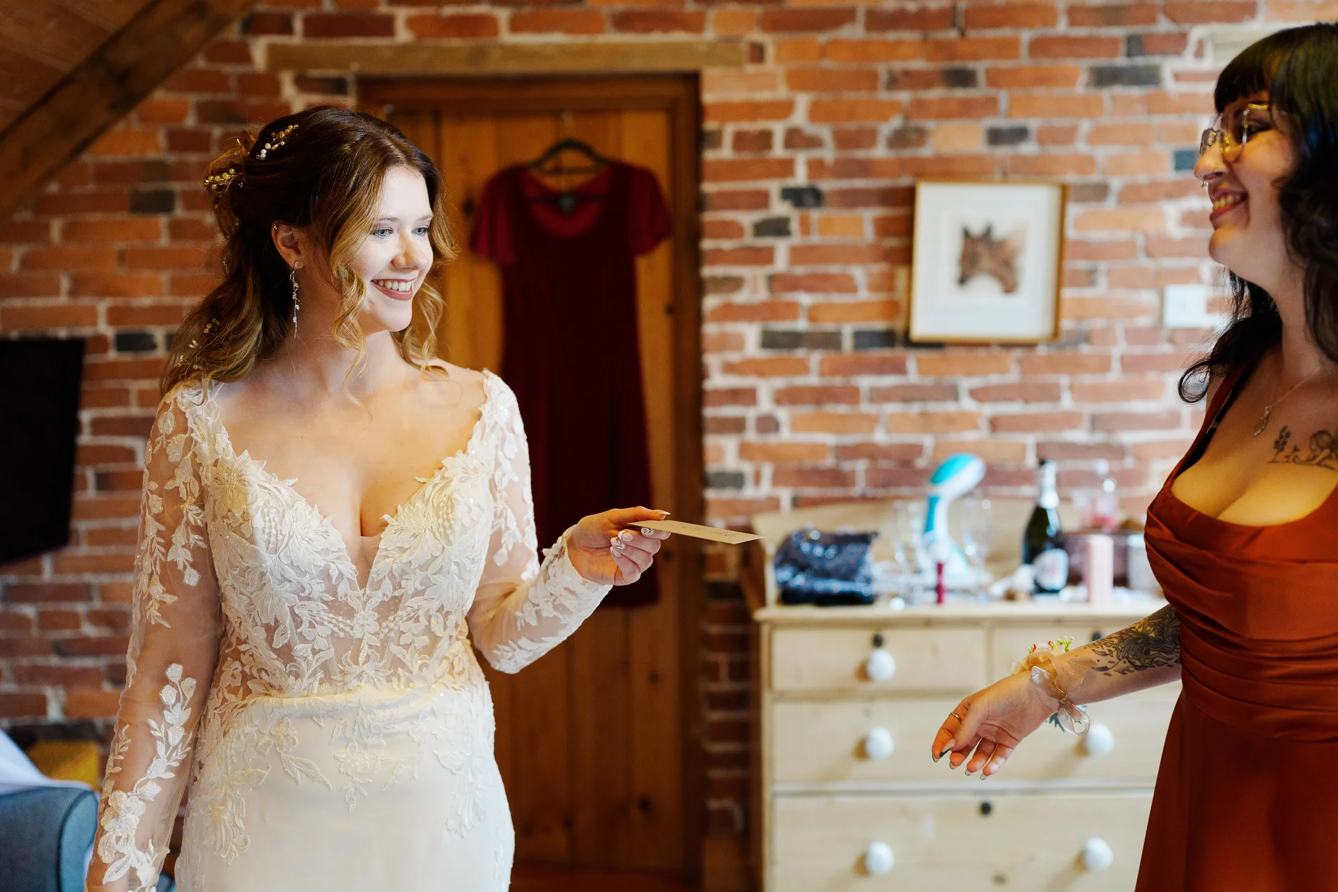 A smiling bride in a long-sleeved lace wedding dress holds a card while reading a letter during morning preparations with her bridesmaid in a rustic brick room.