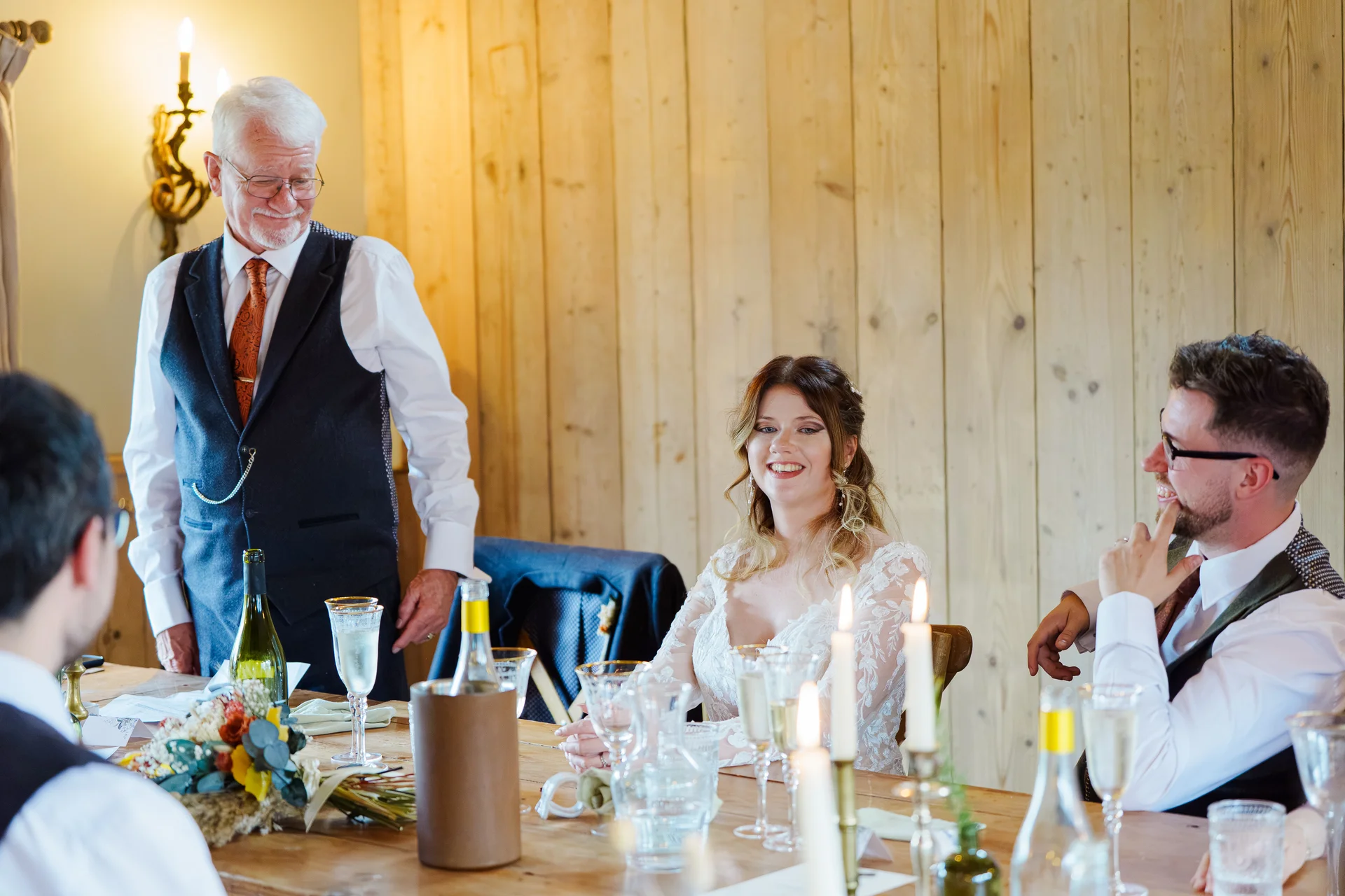 A proud father delivers a speech at the top table while the smiling bride and groom listen and laugh during the wedding breakfast.