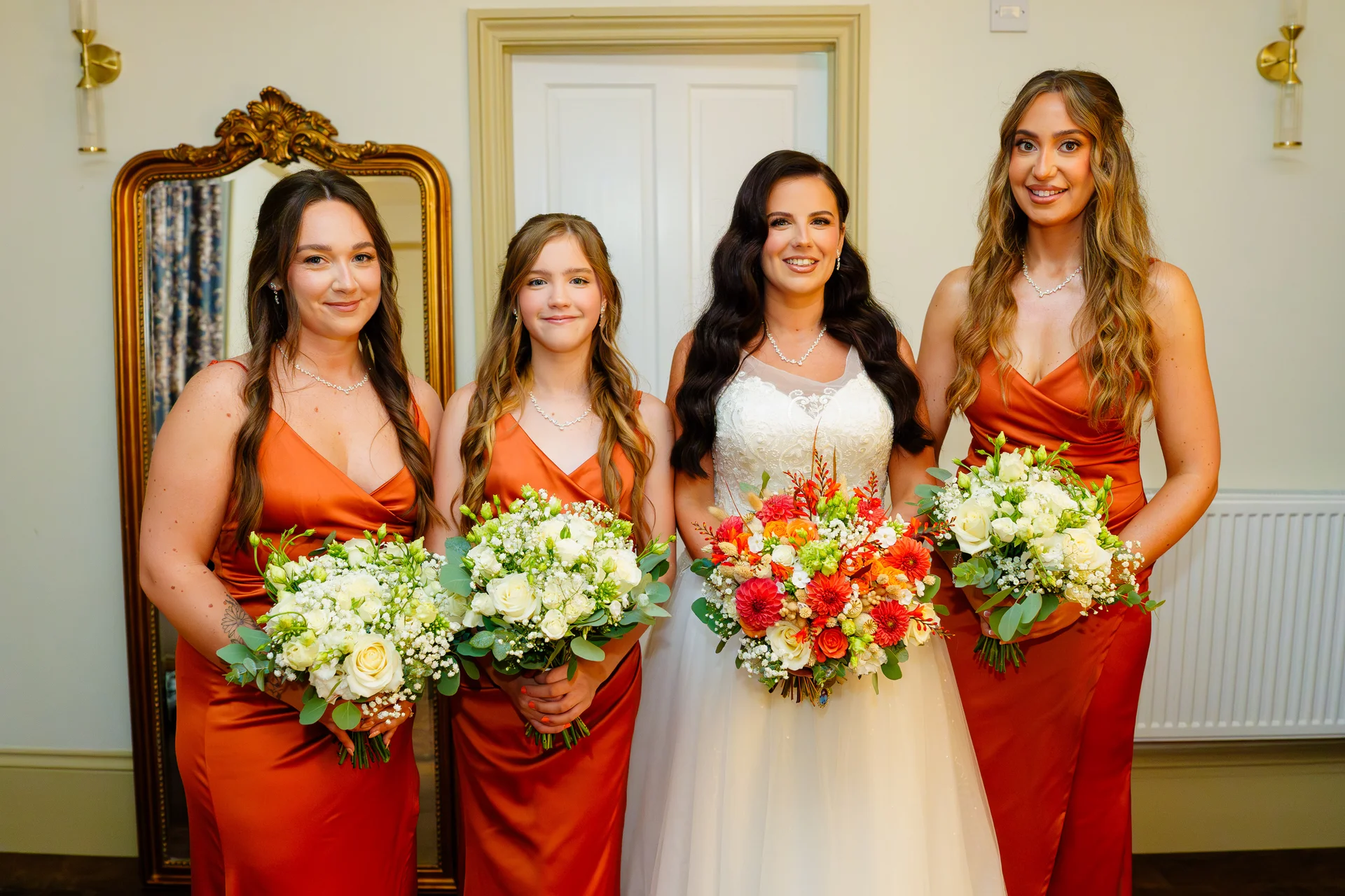 A smiling bride in a white gown and three bridesmaids in satin burnt orange dresses pose together indoors, each holding vibrant floral bouquets.