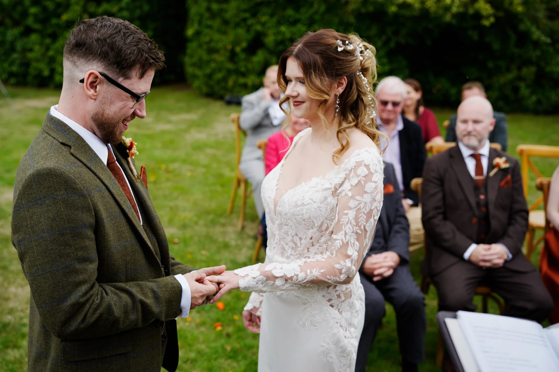 The groom smiles as he gently slides a ring onto the bride's finger during their outdoor ceremony, surrounded by seated guests.