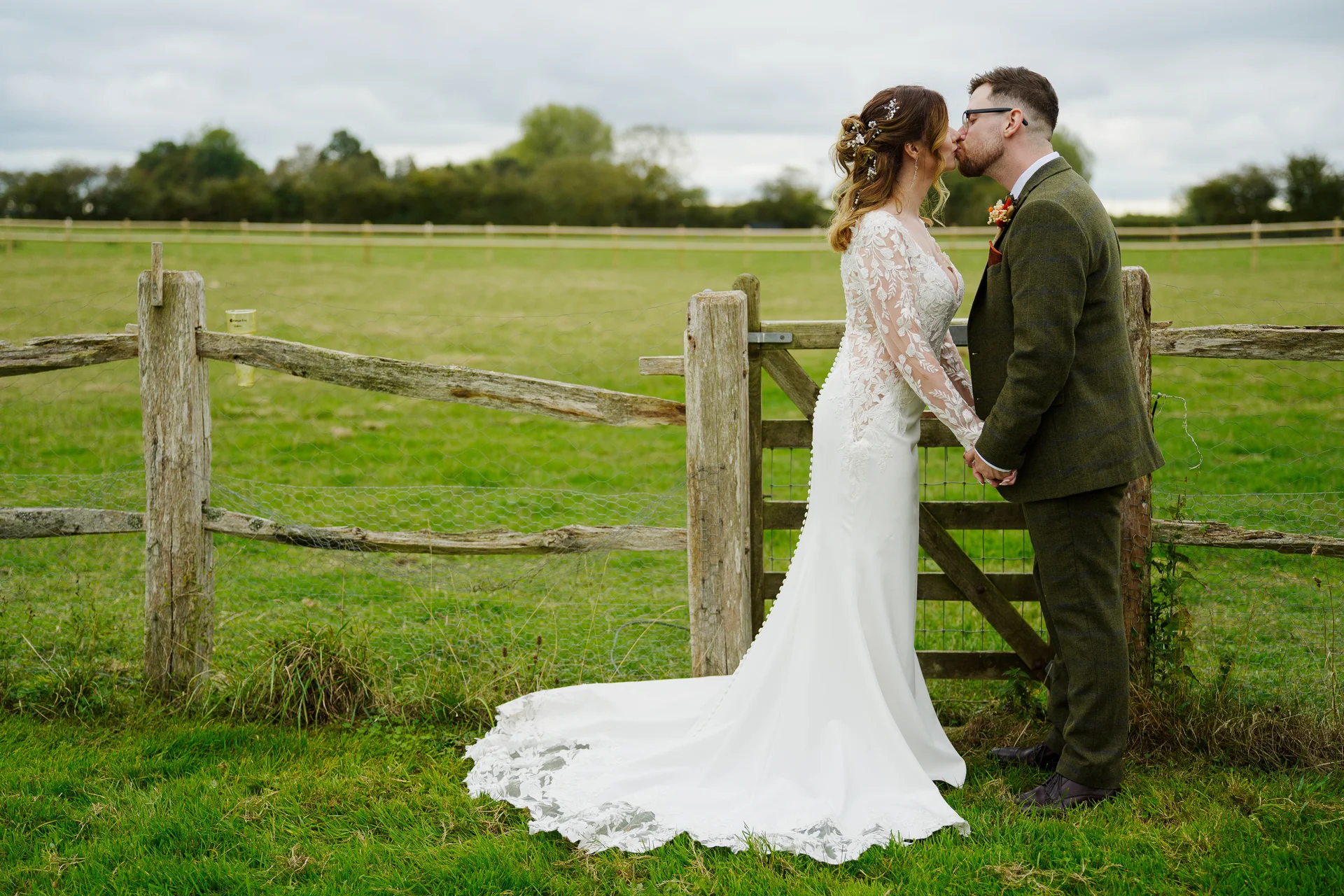 Newlyweds share a tender kiss by a rustic wooden fence in a lush green countryside field on their wedding day.