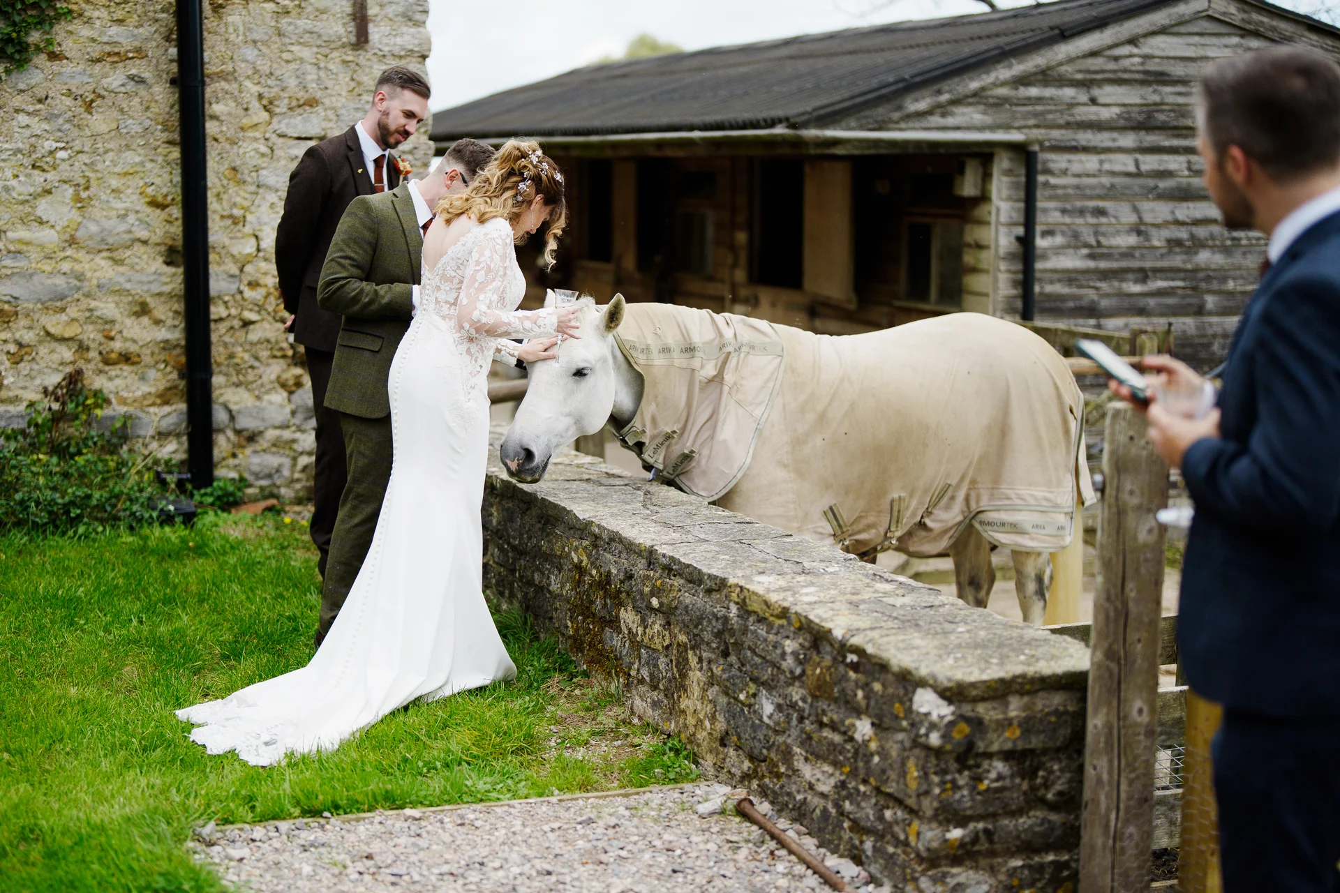 The bride in a long sleeved white gown and groom in a tweed suit interact with a white horse standing by a stone wall at a rustic farm wedding venue.
