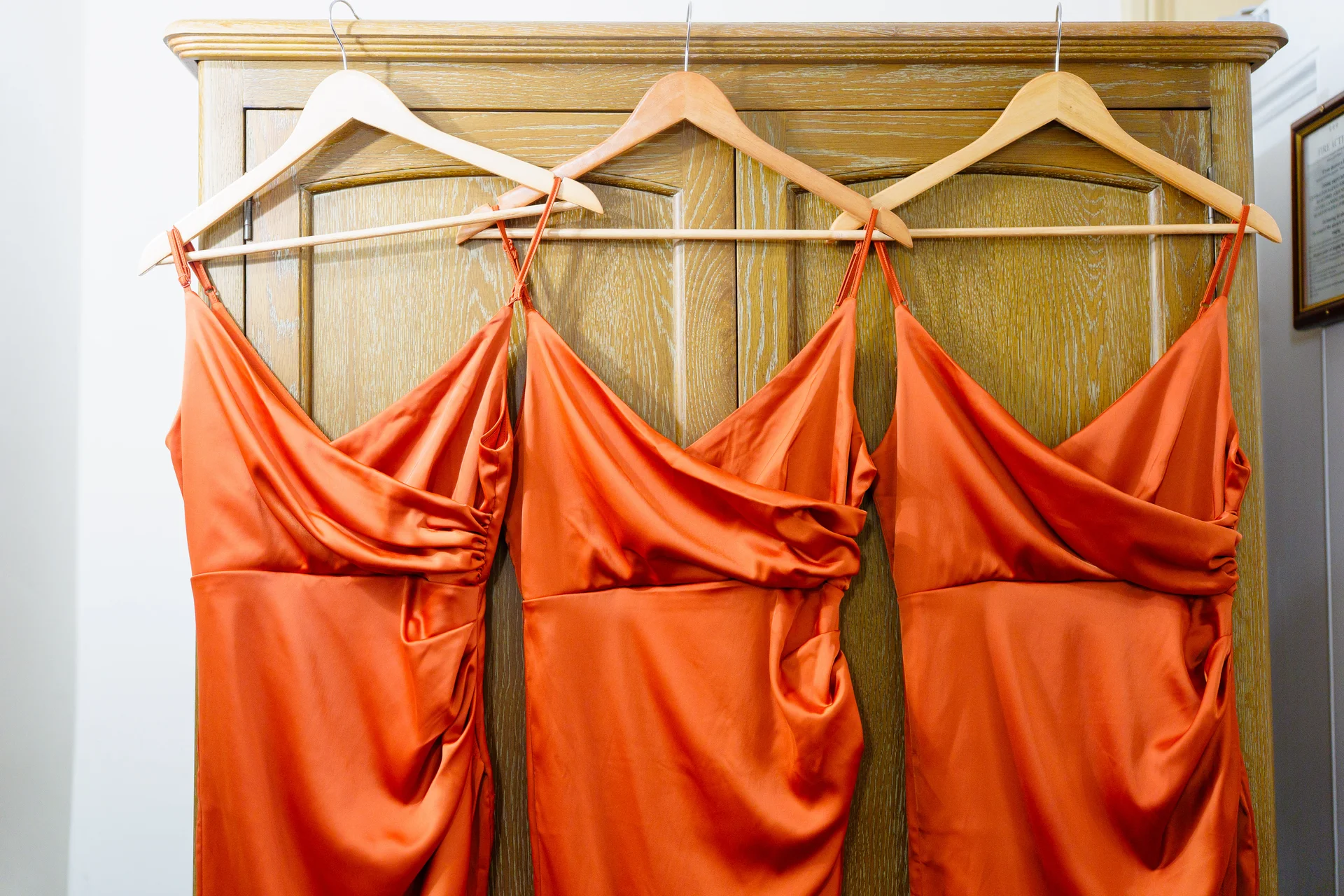 Three elegant burnt orange satin slip dresses with draped bodices hang neatly on wooden coat hangers against a wooden wardrobe during bridal preparations.