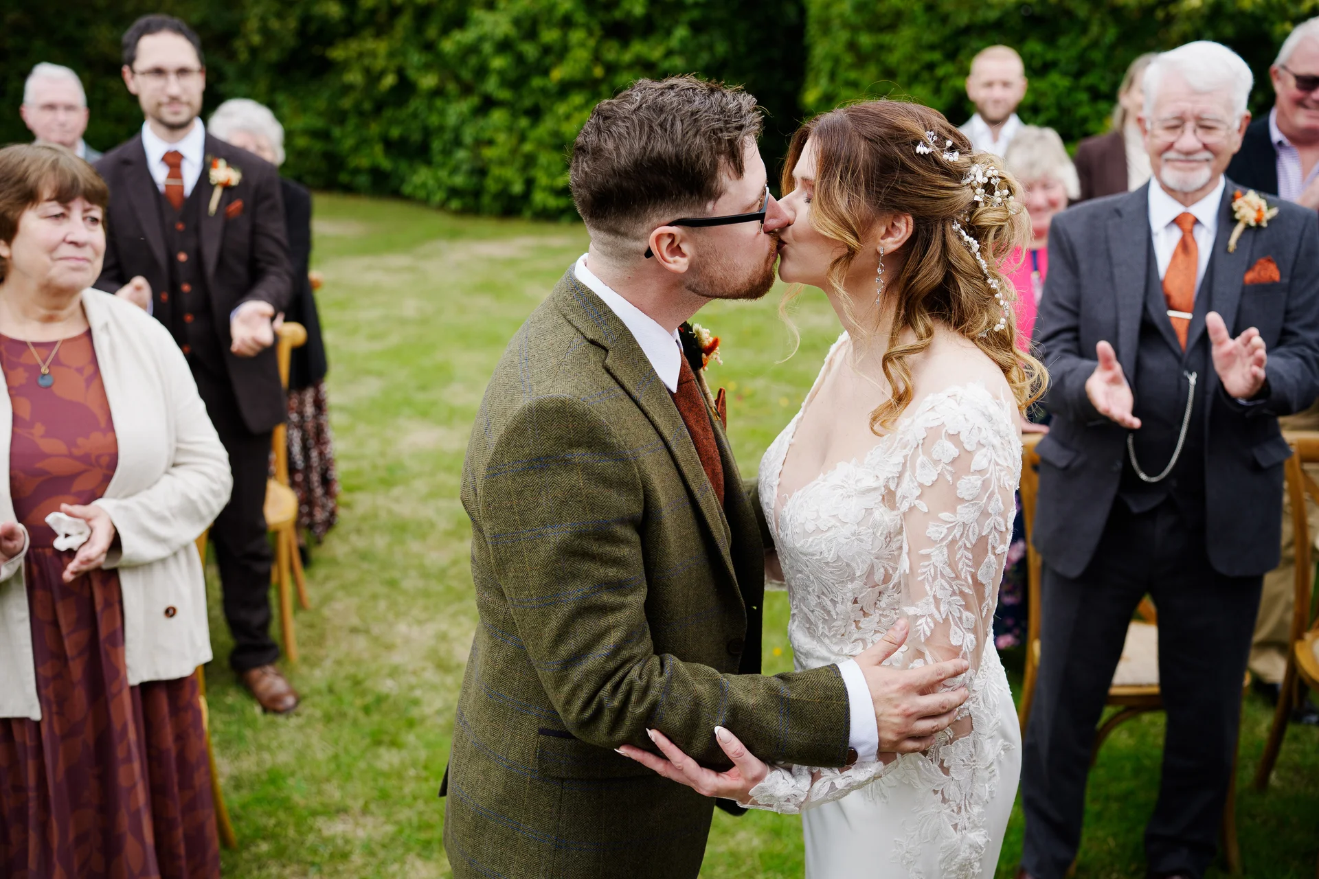 Newlywed bride and groom share their first kiss during an outdoor ceremony while wedding guests applaud in the background.