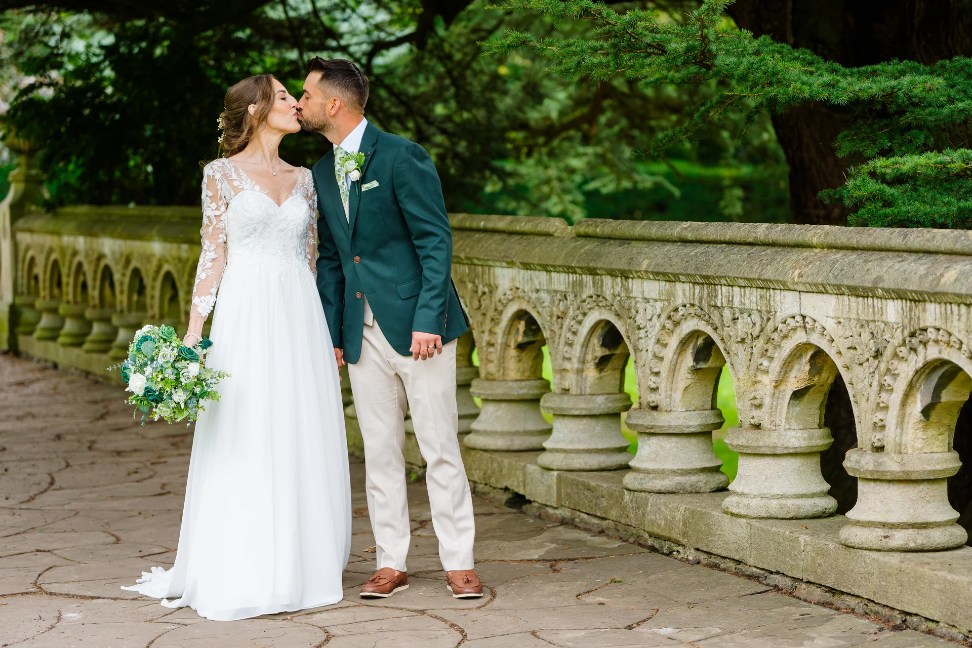 A newlywed couple shares a tender kiss on a paved stone bridge with decorative balustrades, surrounded by lush green trees.