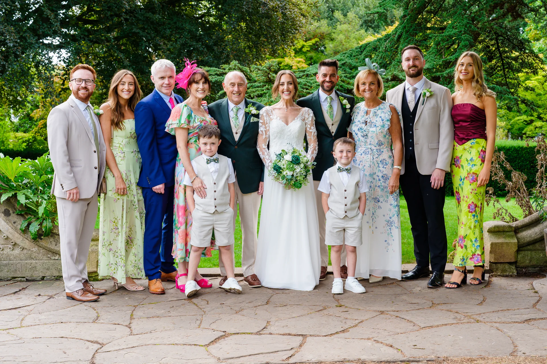 A large wedding party and family group pose smiling together on a paved terrace surrounded by lush green trees and gardens.