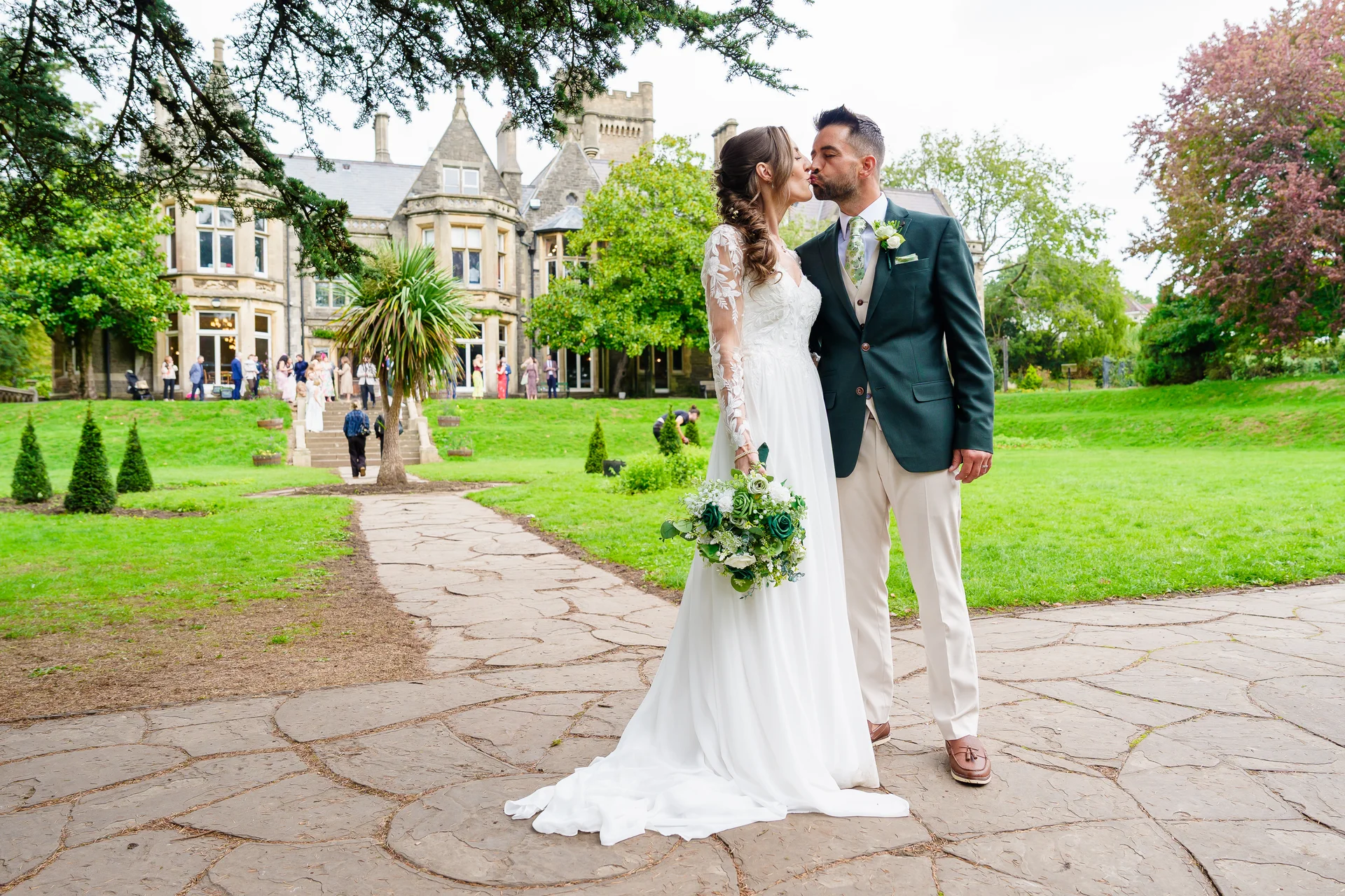 A newlywed couple shares a tender kiss on a stone pathway in front of a grand country house with lush green lawns.