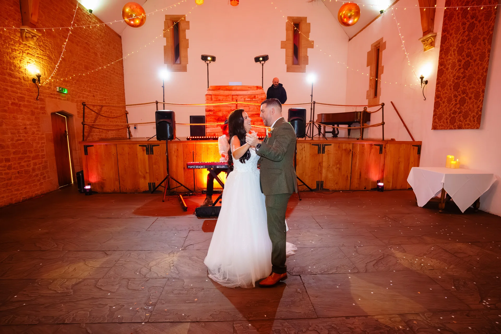 A newlywed couple share a joyful first dance together in a rustic stone barn venue decorated with warm string lights.