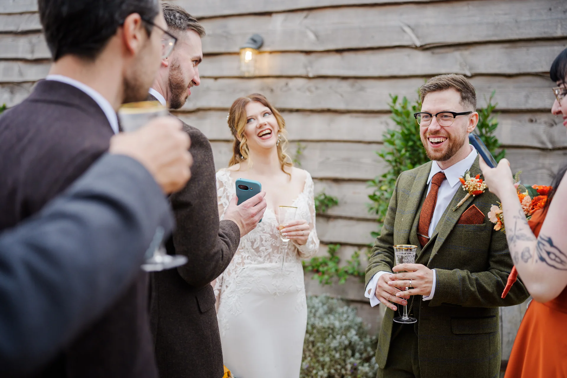 A cheerful group of wedding guests and the smiling bride and groom mingling outside against a wooden barn wall while holding drinks.