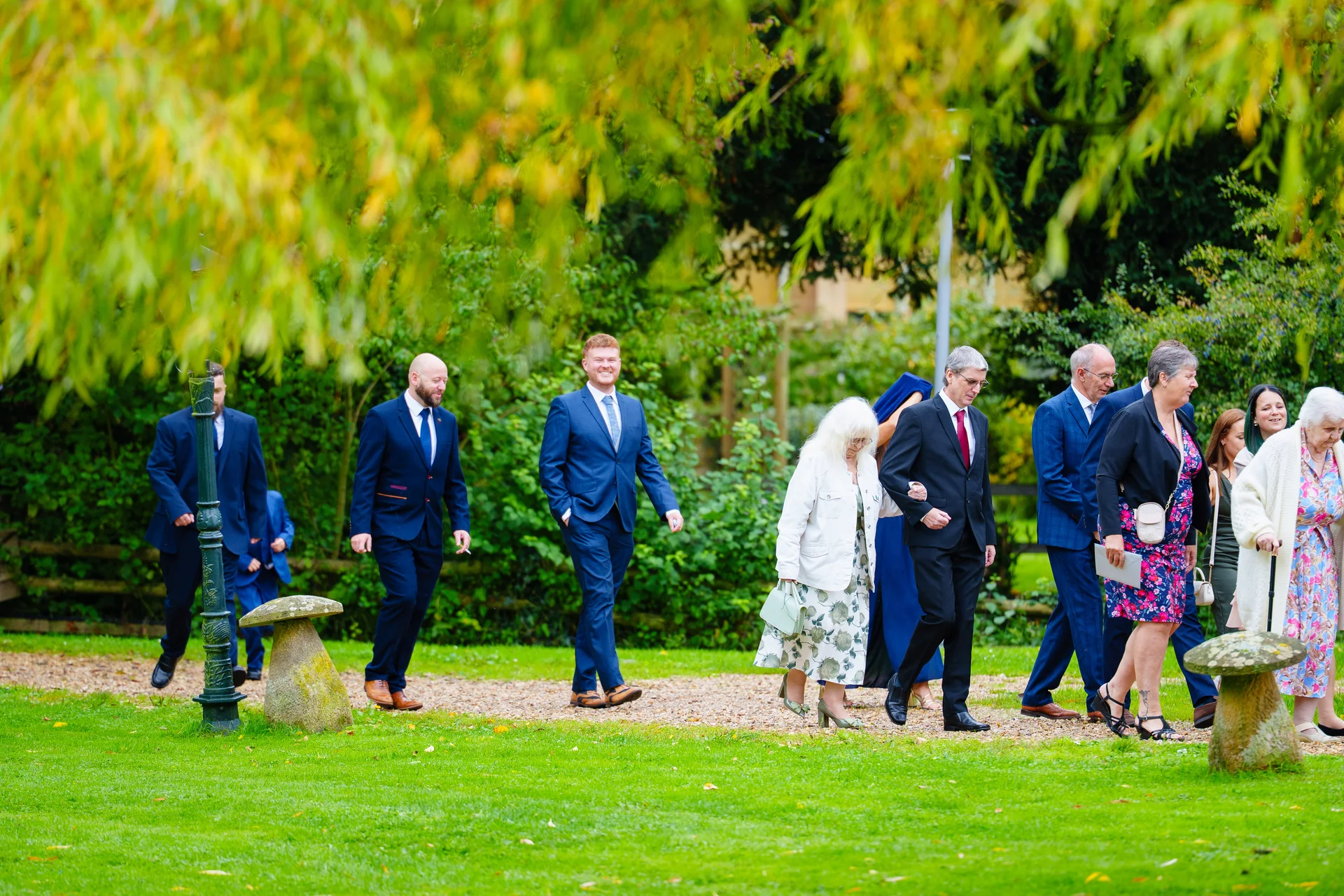 Wedding guests in smart suits and dresses walk along a gravel path through lush green gardens, framed by overhanging golden tree foliage.