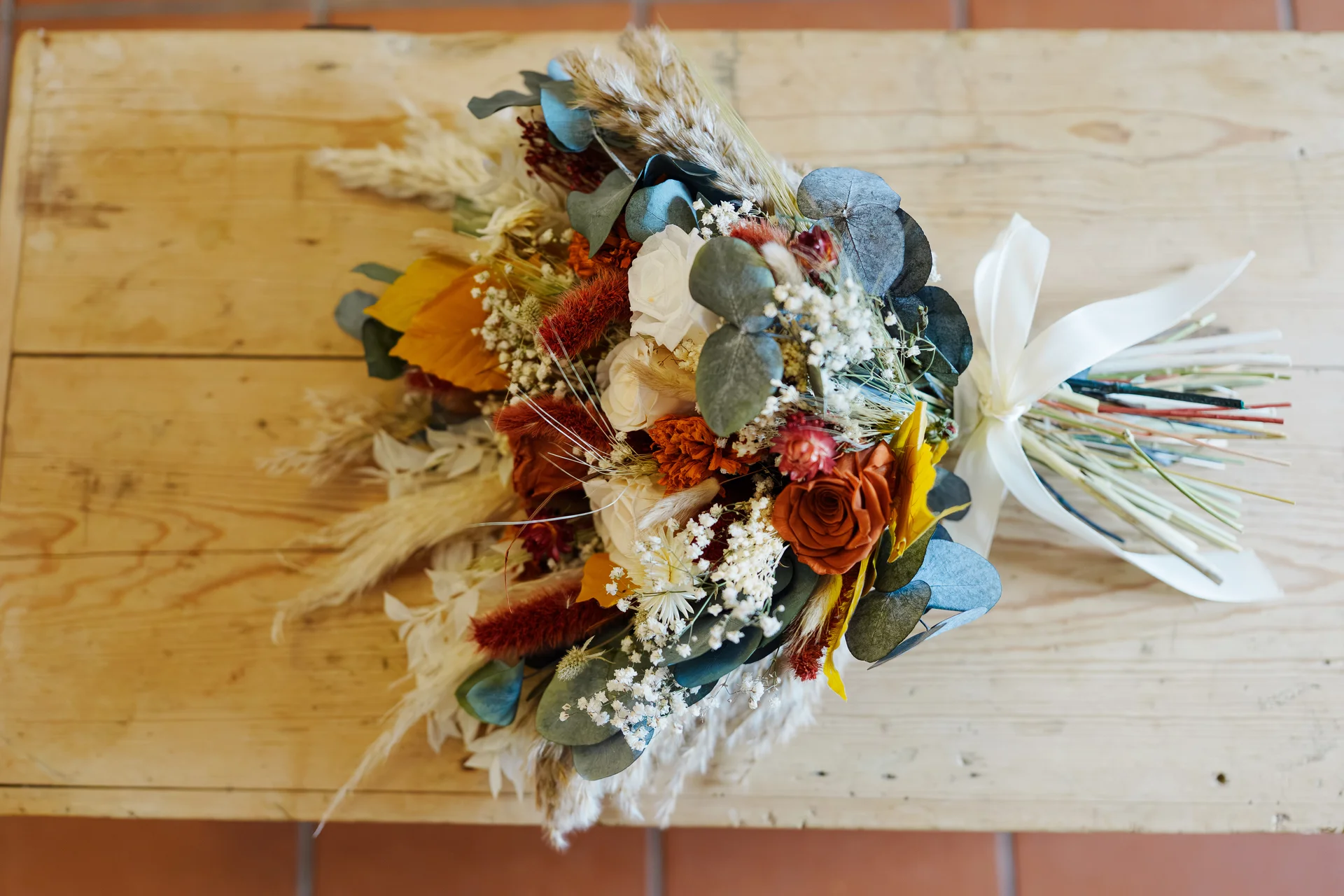 A stunning autumnal bridal bouquet featuring dried flowers, burnt orange roses, and eucalyptus rests on a rustic wooden surface.