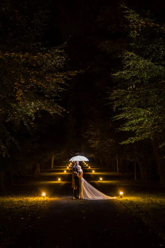 A newlywed couple embraces under a glowing umbrella on a dark, tree-lined path illuminated by warm ground lights at night.
