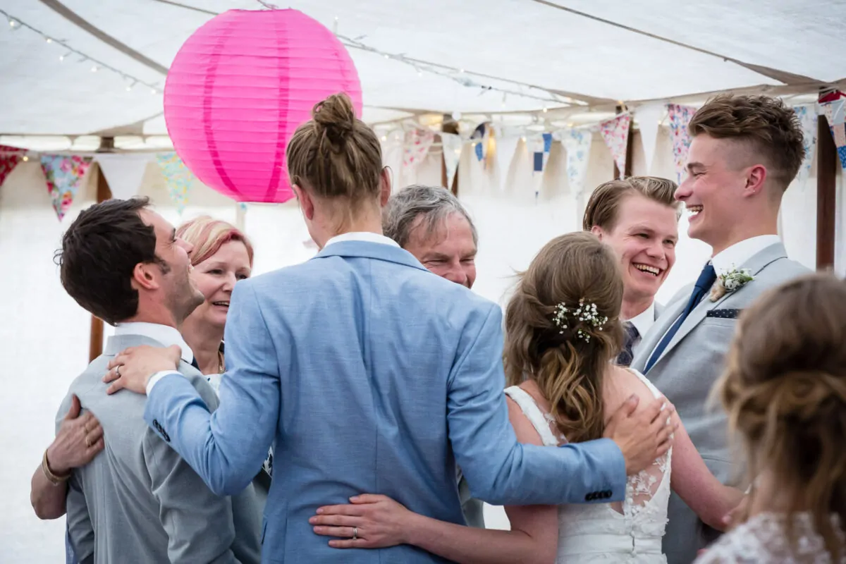 A joyful group of wedding guests share a warm embrace and share a laugh under a marquee decorated with bunting and paper lanterns.