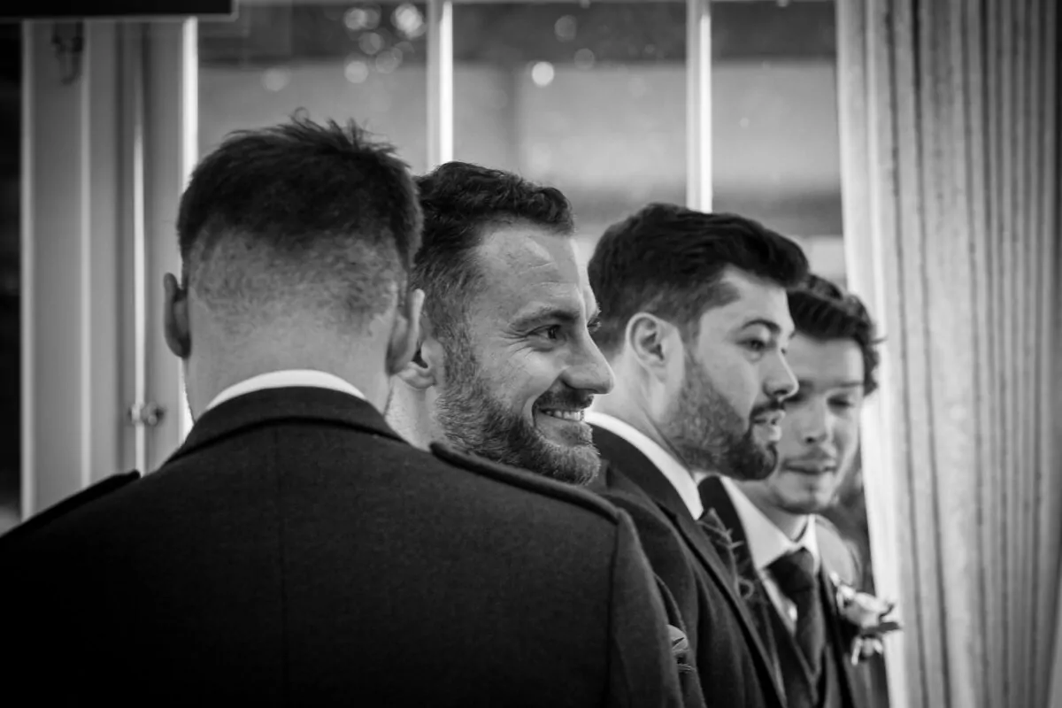 A candid black and white shot captures a line of smiling groomsmen standing near a window, dressed in smart dark suits.