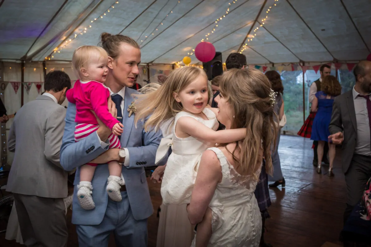 A newlywed couple dancing inside a marquee adorned with fairy lights, happily holding two young children while guests mingle in the background.