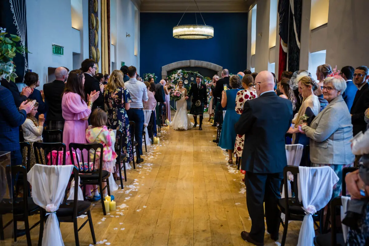Newlyweds walk down the aisle hand in hand, smiling at their cheering guests as white petals and candles decorate the wooden floor.
