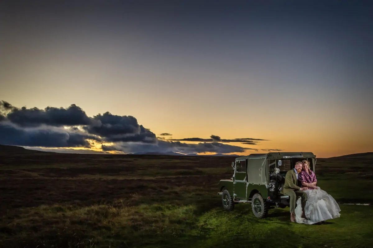 A newlywed couple sits on the back of a vintage green land rover amidst an expansive moorland landscape under a dramatic sunset sky.